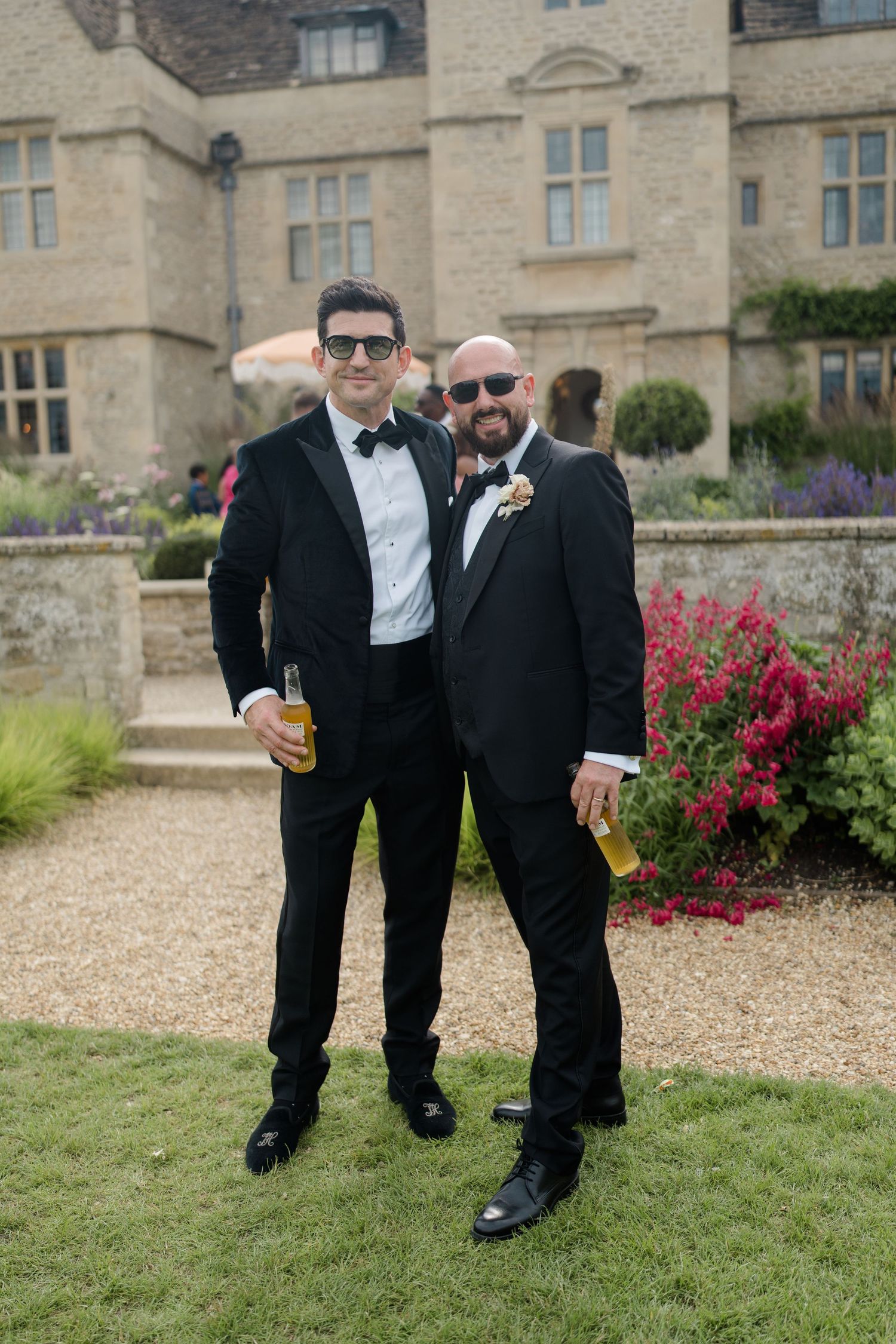 Two male wedding guests in black tuxedos with bow ties and sunglasses posing with beer bottles at manor house wedding.