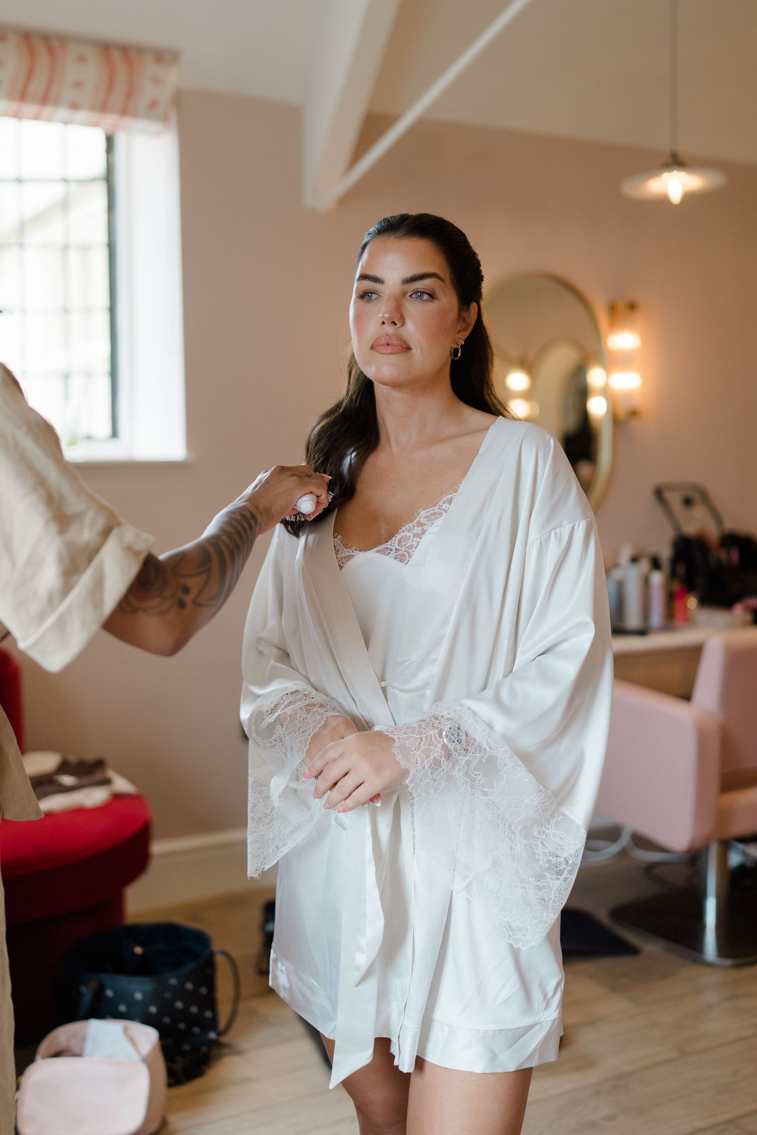 A bride in a white lace robe having her hair styled in a bright salon with mirrors.