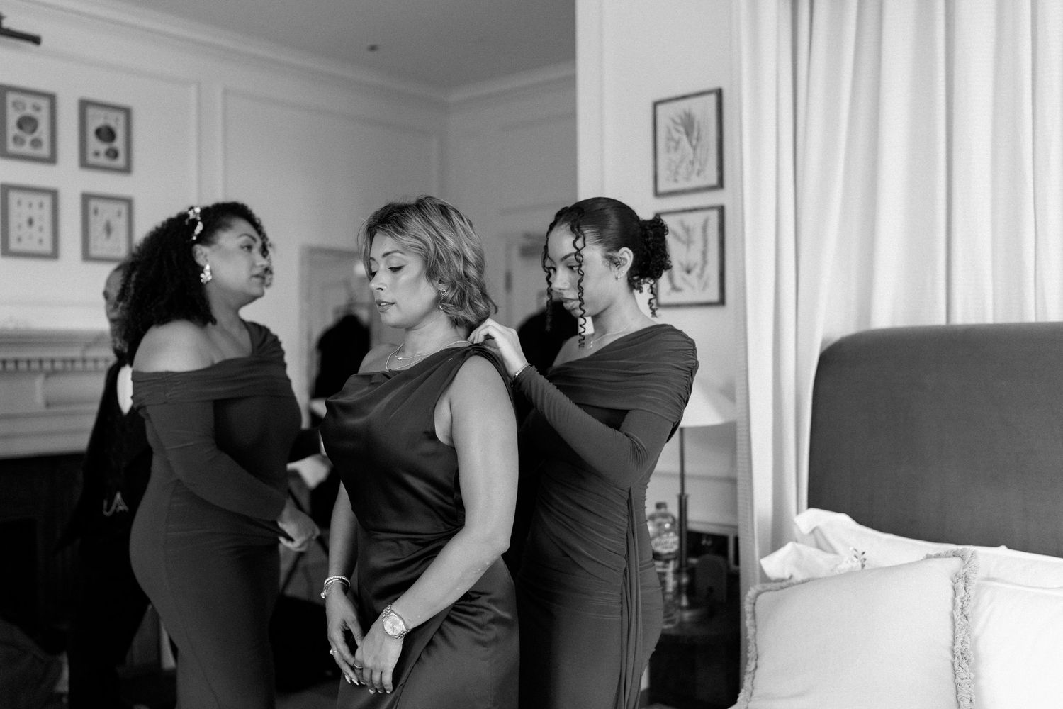 Bridesmaids in dark green dresses helping each other prepare in elegant bedroom, black and white.