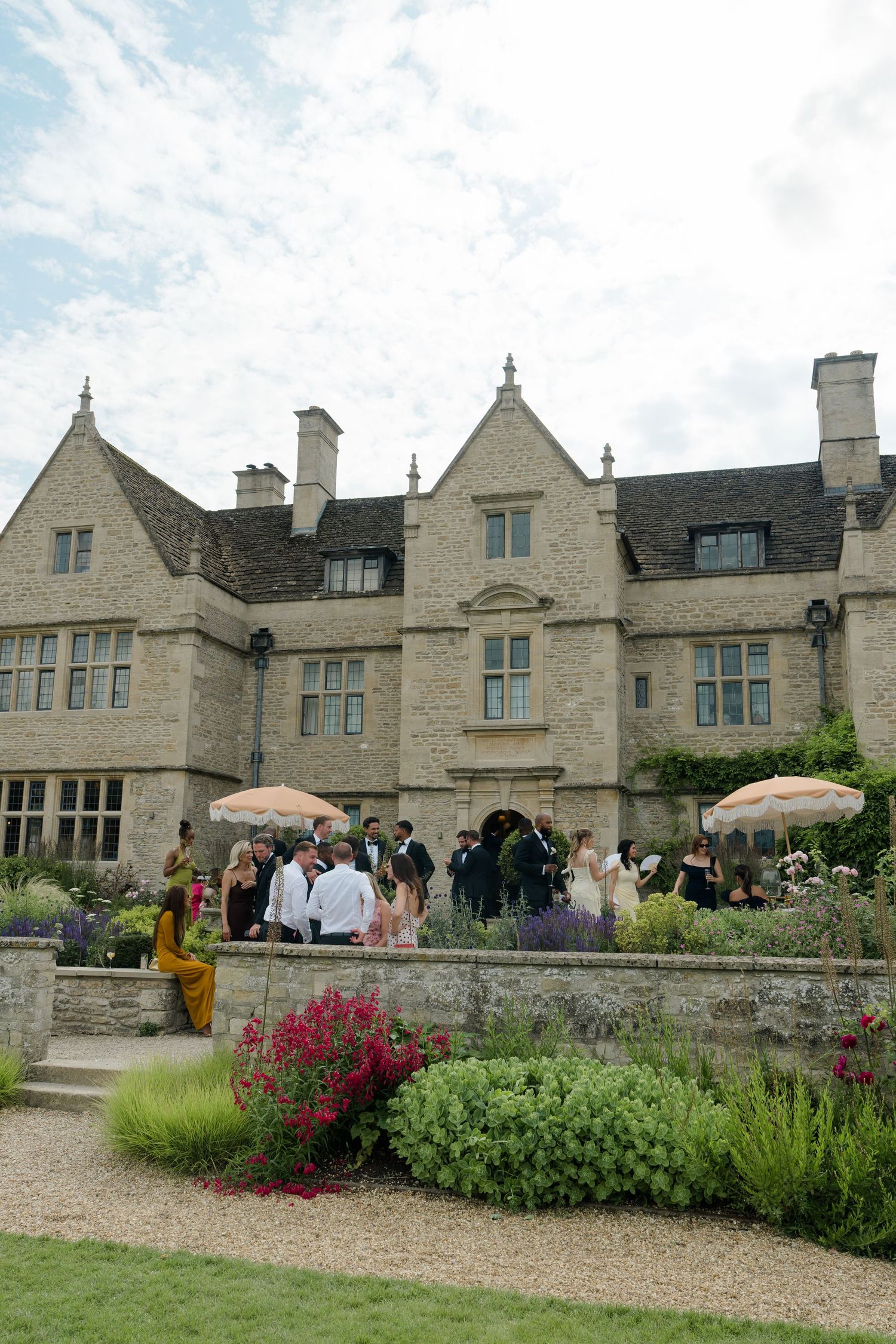 Wedding guests mingling outside historic Cotswolds stone manor house with garden flowers.