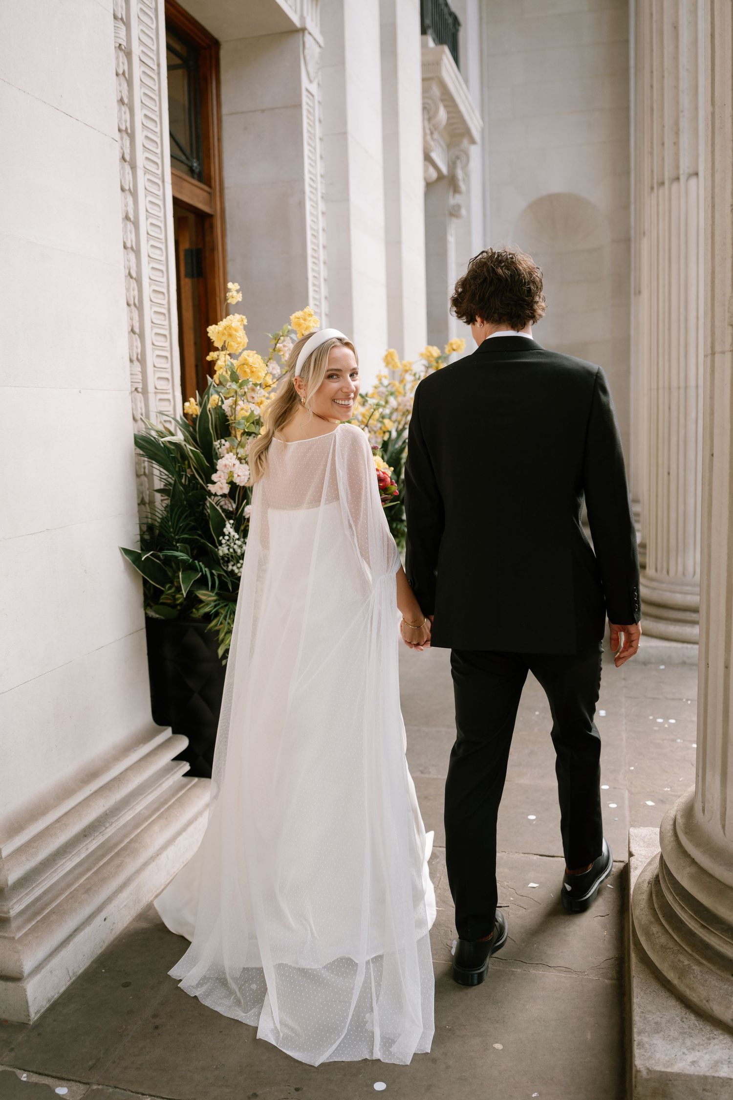 A bride in flowing white dress and groom walking past ornate columns with yellow flowers.