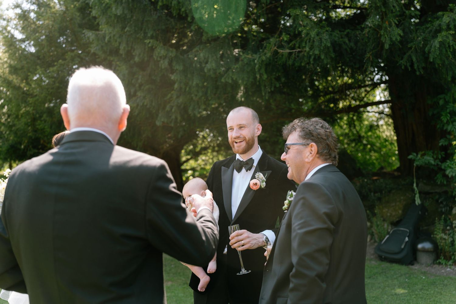 Wedding guests chat during an outdoor reception as sunlight filters through the trees.