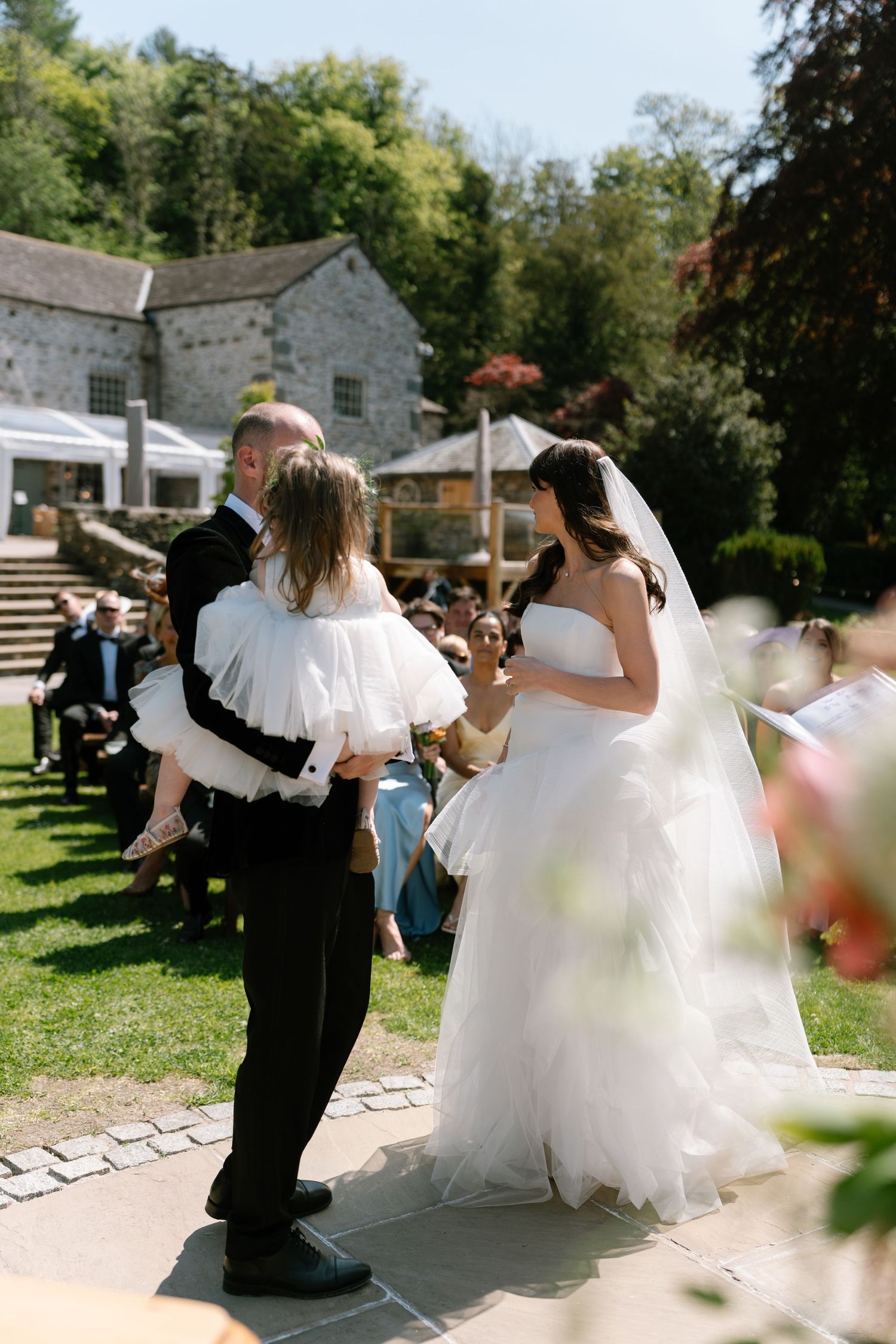 Wedding celebration on a sunny day with guests dancing on a stone patio near historic buildings.