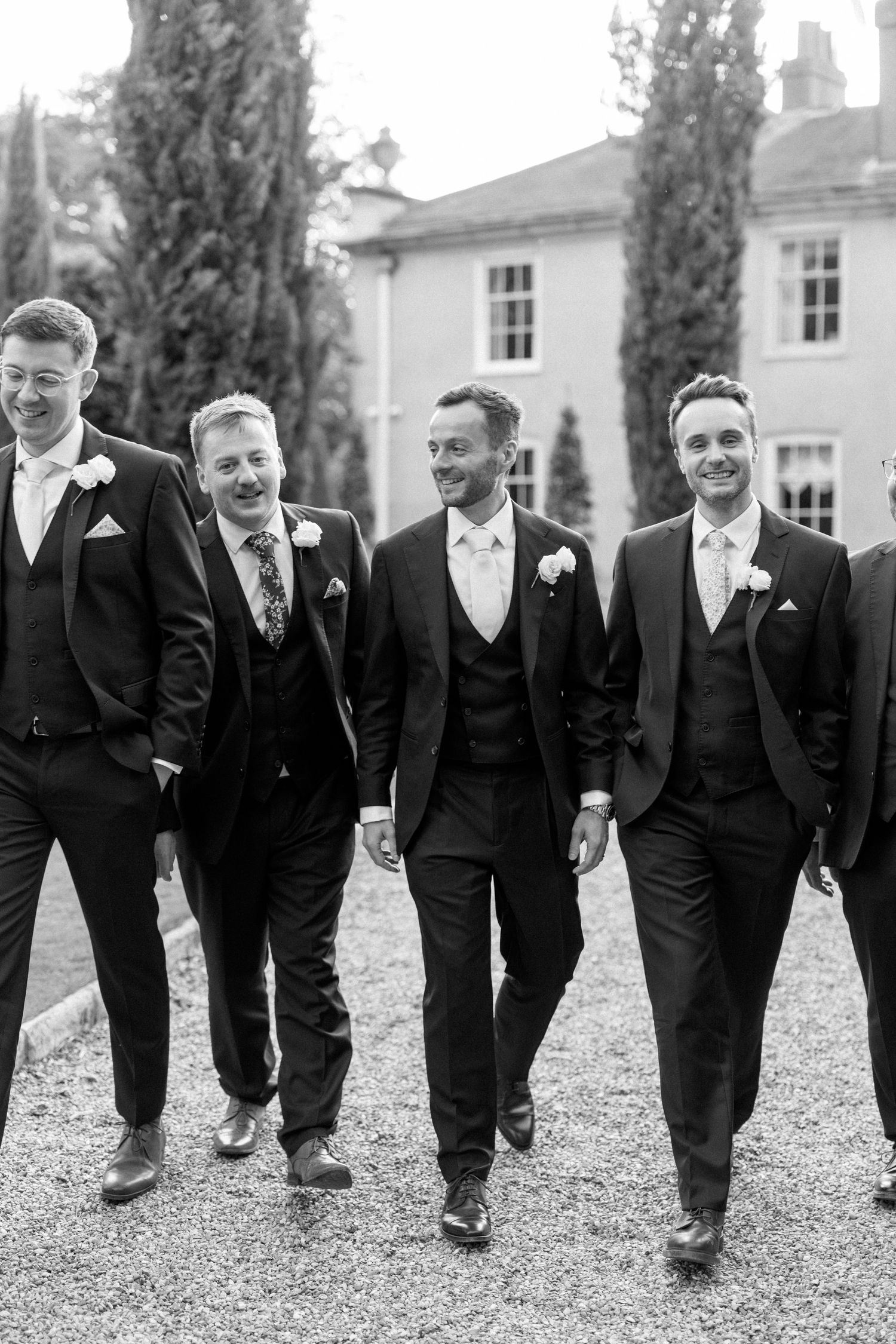 Four groomsmen walking and smiling on a gravel path in black and white photography.