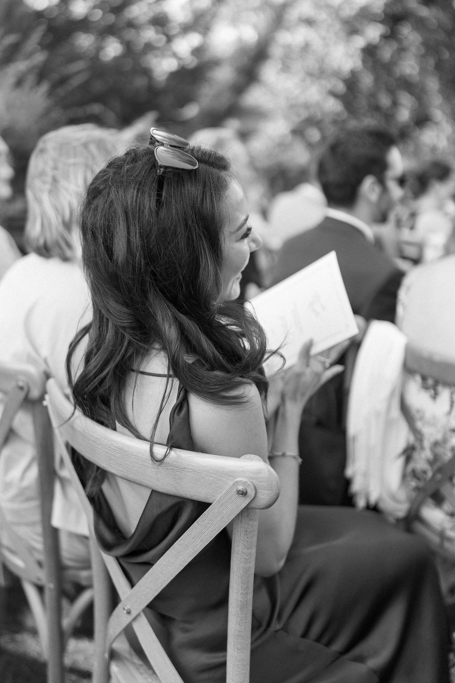 Guest in off-shoulder dress watches wedding ceremony from wooden chair.