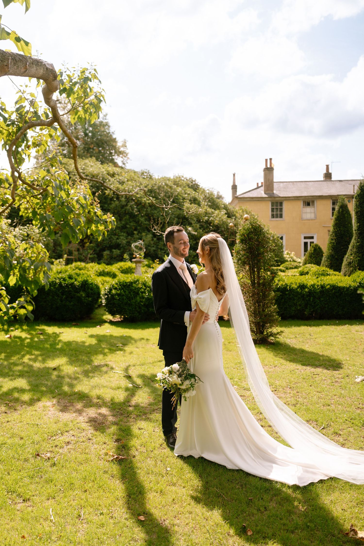 Sunlit wedding photos in a lush garden setting with historic manor house visible.