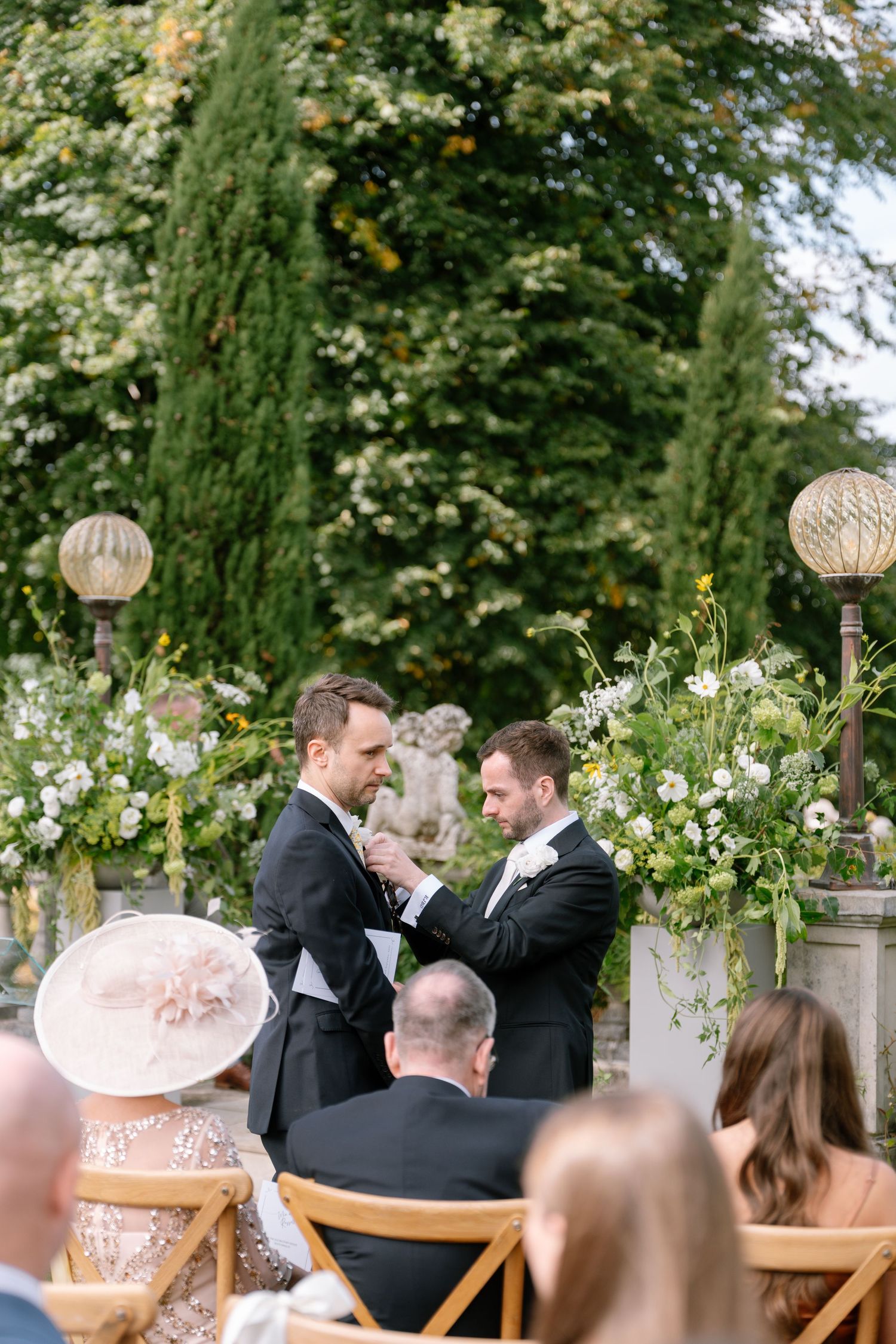 Wedding ceremony guests seated in wooden chairs with hanging lantern decorations in a garden setting.