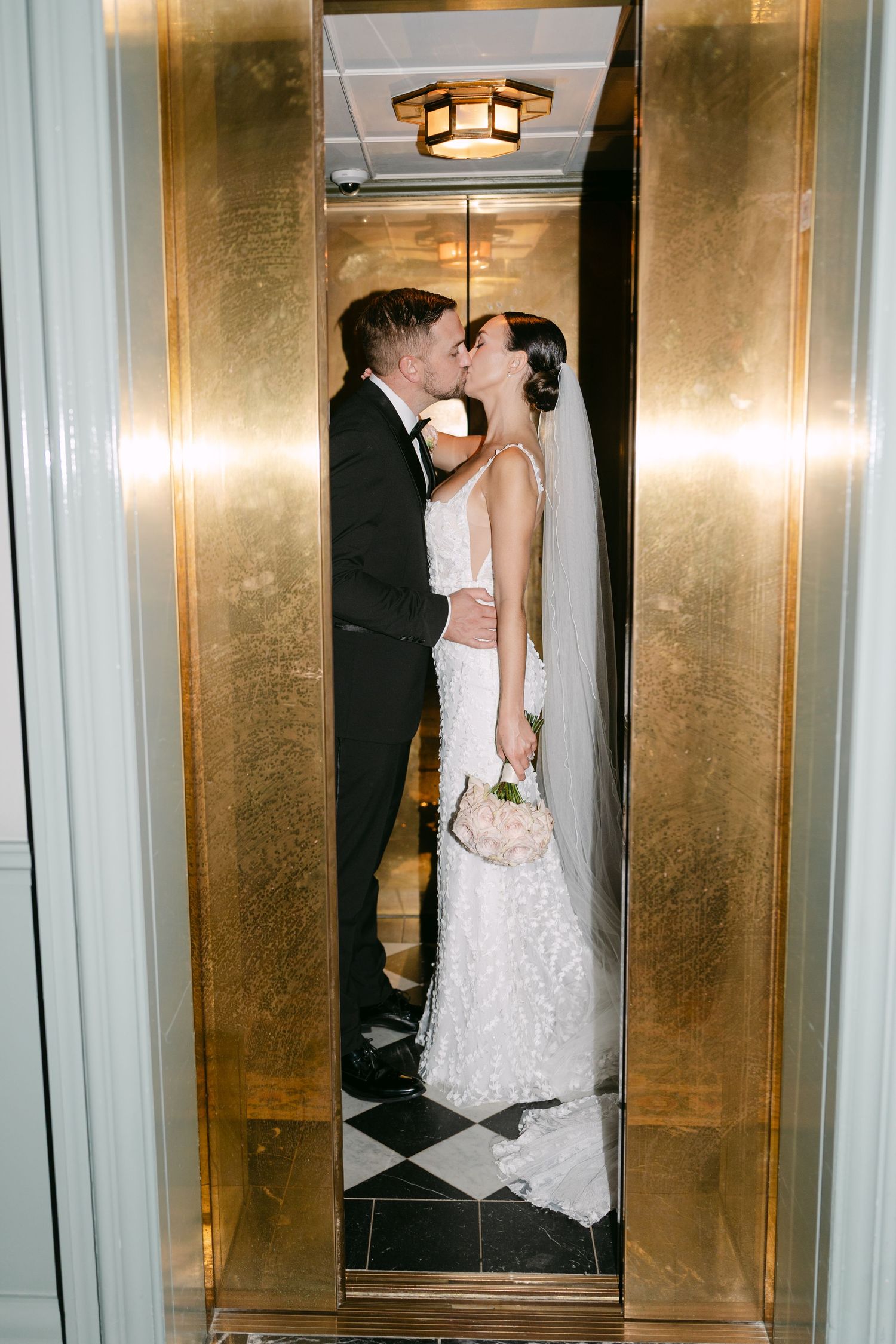 A wedding couple embraces in a luxurious gold-paneled elevator with checkered floor.