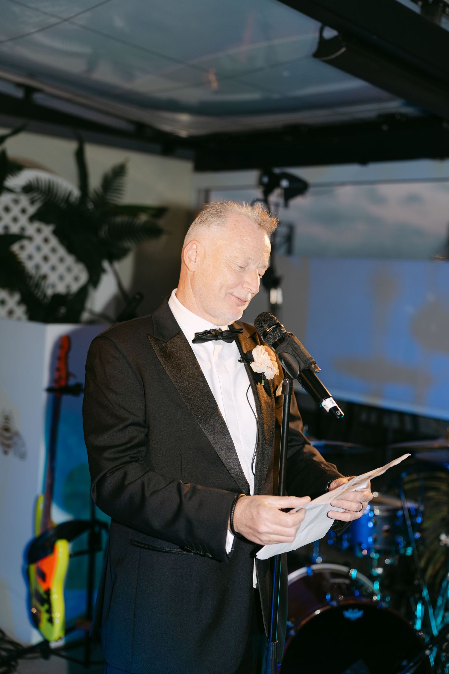 Man in tuxedo giving a speech at wedding reception with band equipment visible in background.