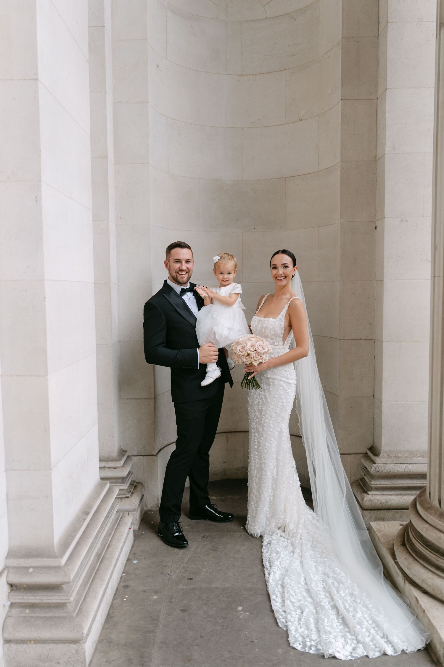 A wedding couple and small child pose between elegant white columns in formal attire.