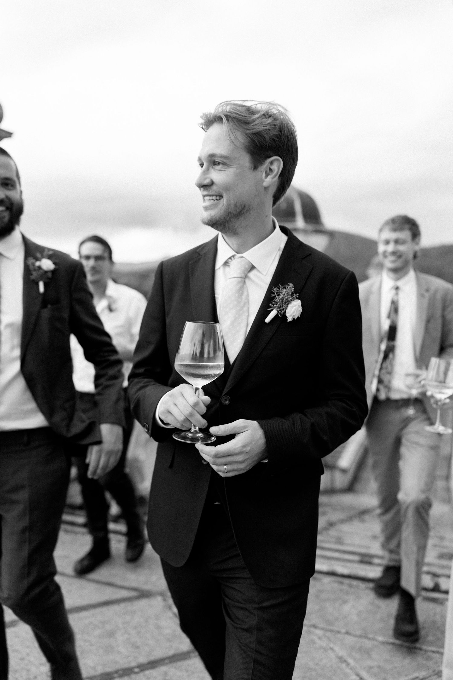 Groomsmen in black suits laugh and socialize during a wedding celebration.