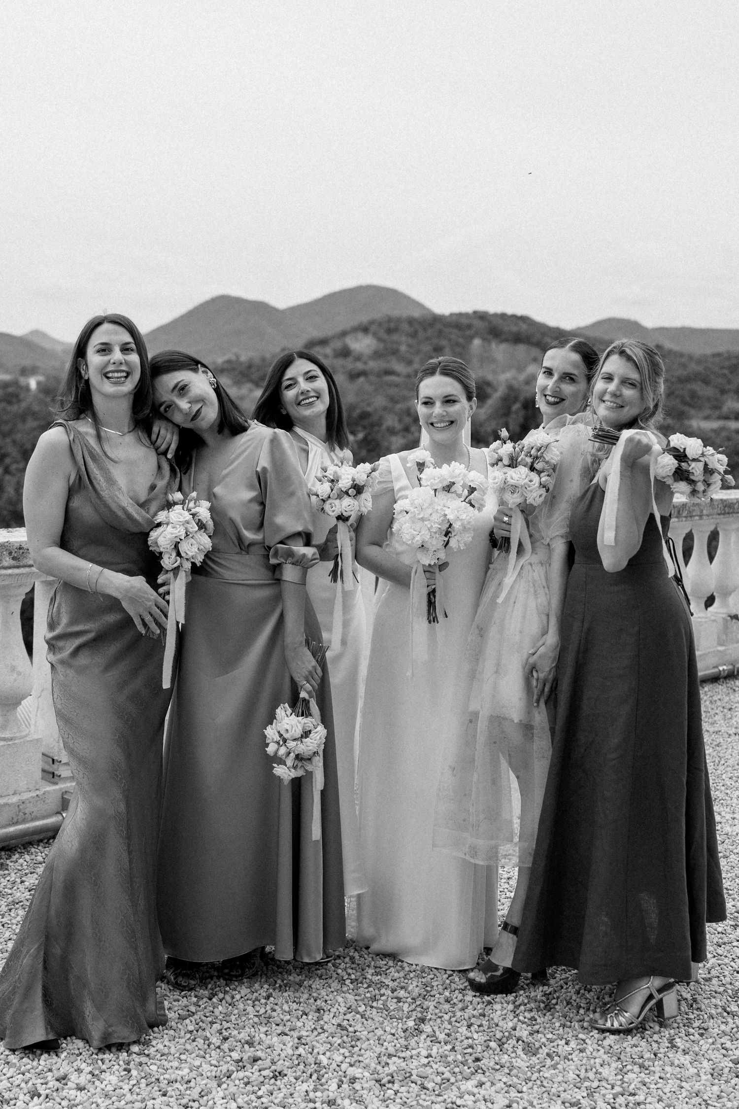 Wedding party in elegant floor-length dresses poses together against a mountain backdrop.