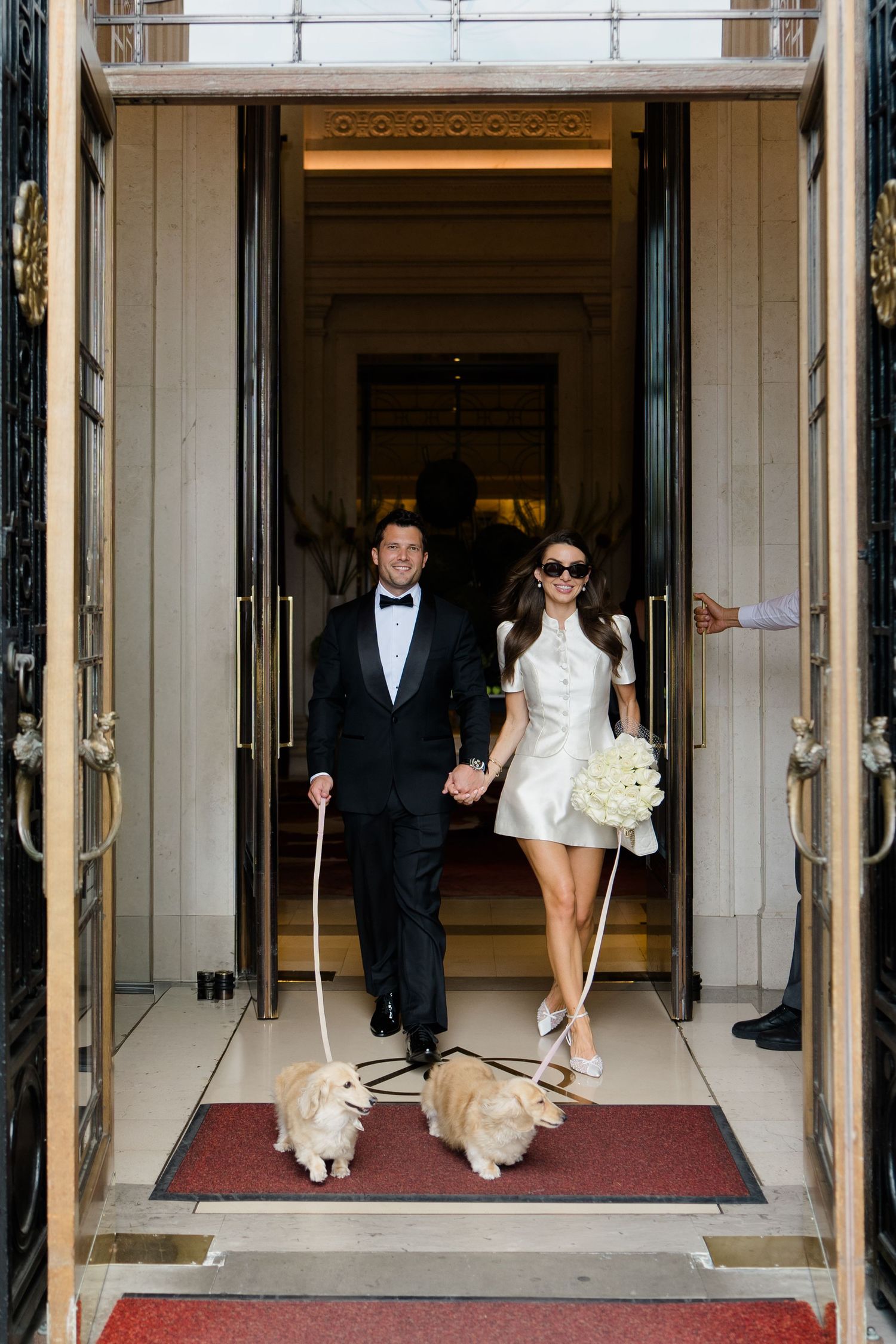 Bride and groom exit elegant venue holding hands, walking two Corgi dogs, bride in mini dress with bouquet, groom in tuxedo.