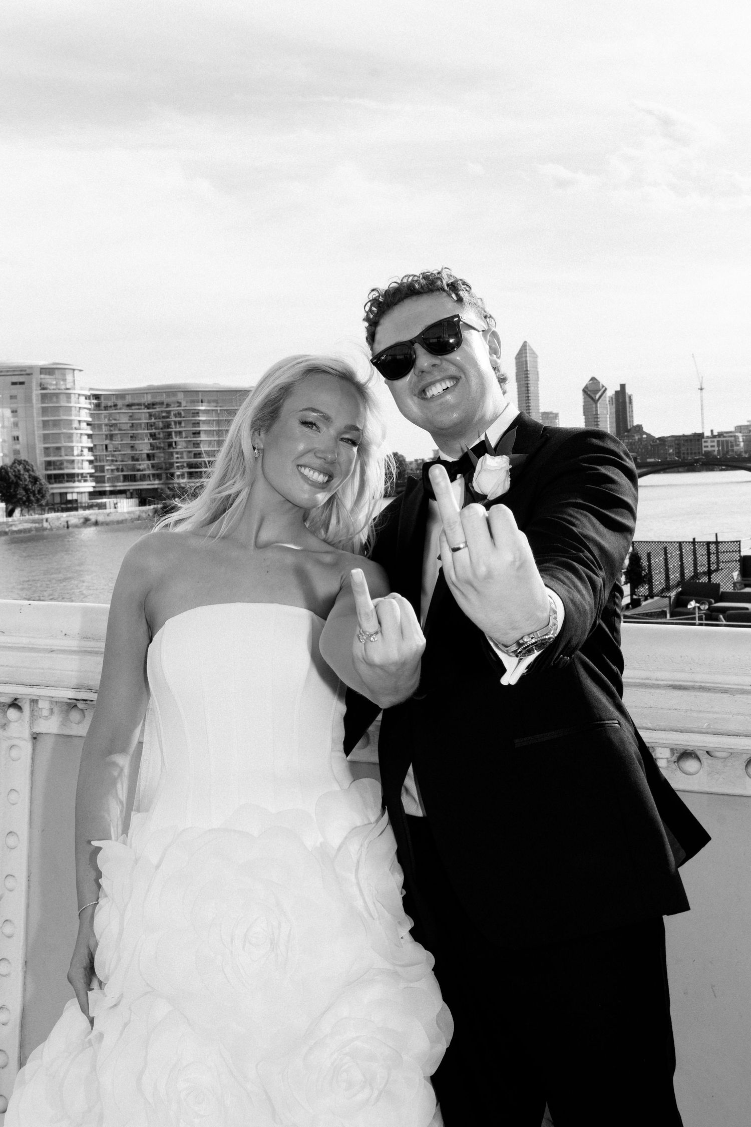Black and white photo of smiling bride and groom showing wedding rings on bridge with city skyline backdrop.