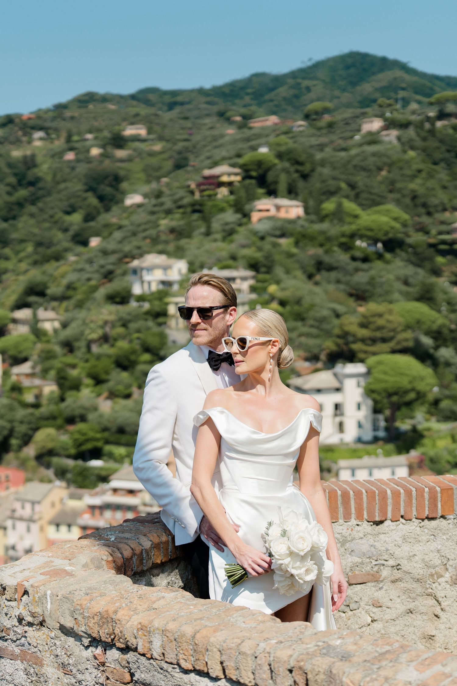 Stylish bride and groom in sunglasses pose on stone wall with lush Italian hillside village backdrop.
