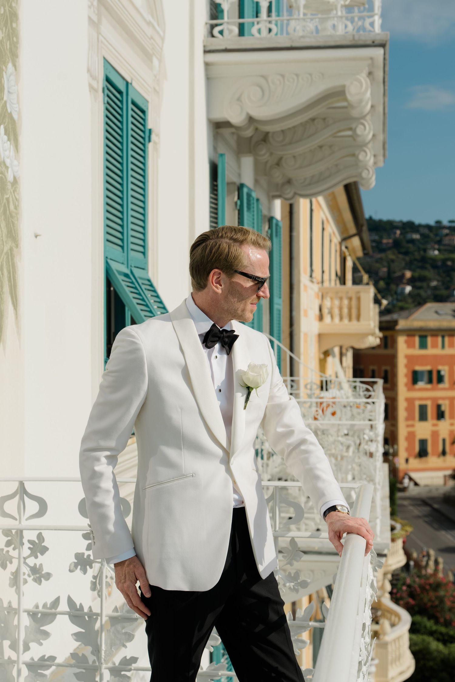 Groom in white tuxedo jacket with black bow tie and boutonniere on ornate balcony in Italian town.