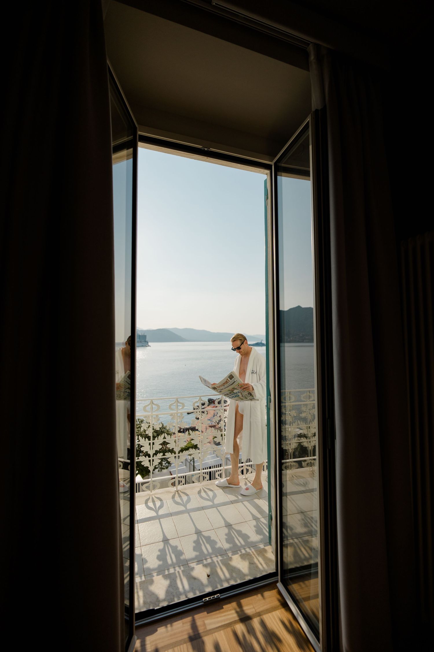Woman in white dress on balcony overlooking scenic sea and mountains, viewed through open glass doors.