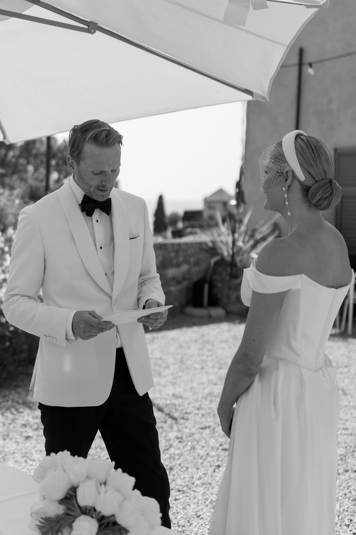 Black and white photo of a groom in white tuxedo reading vows to his bride at an outdoor ceremony.