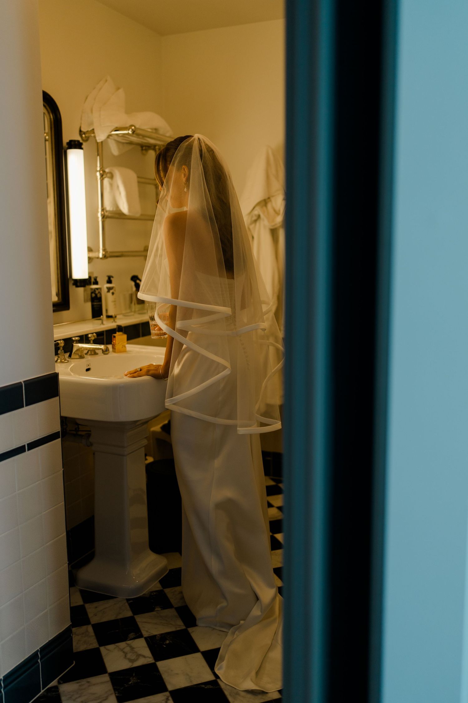 Bride in white wedding dress and veil standing at bathroom sink in black and white tiled room.