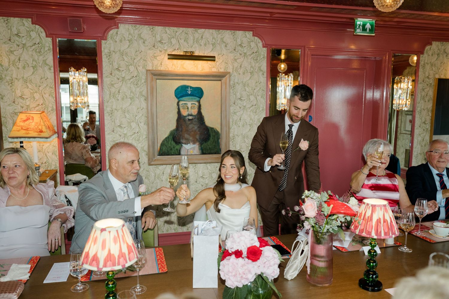 Groom gives wedding speech at reception table while bride and guests listen and smile.