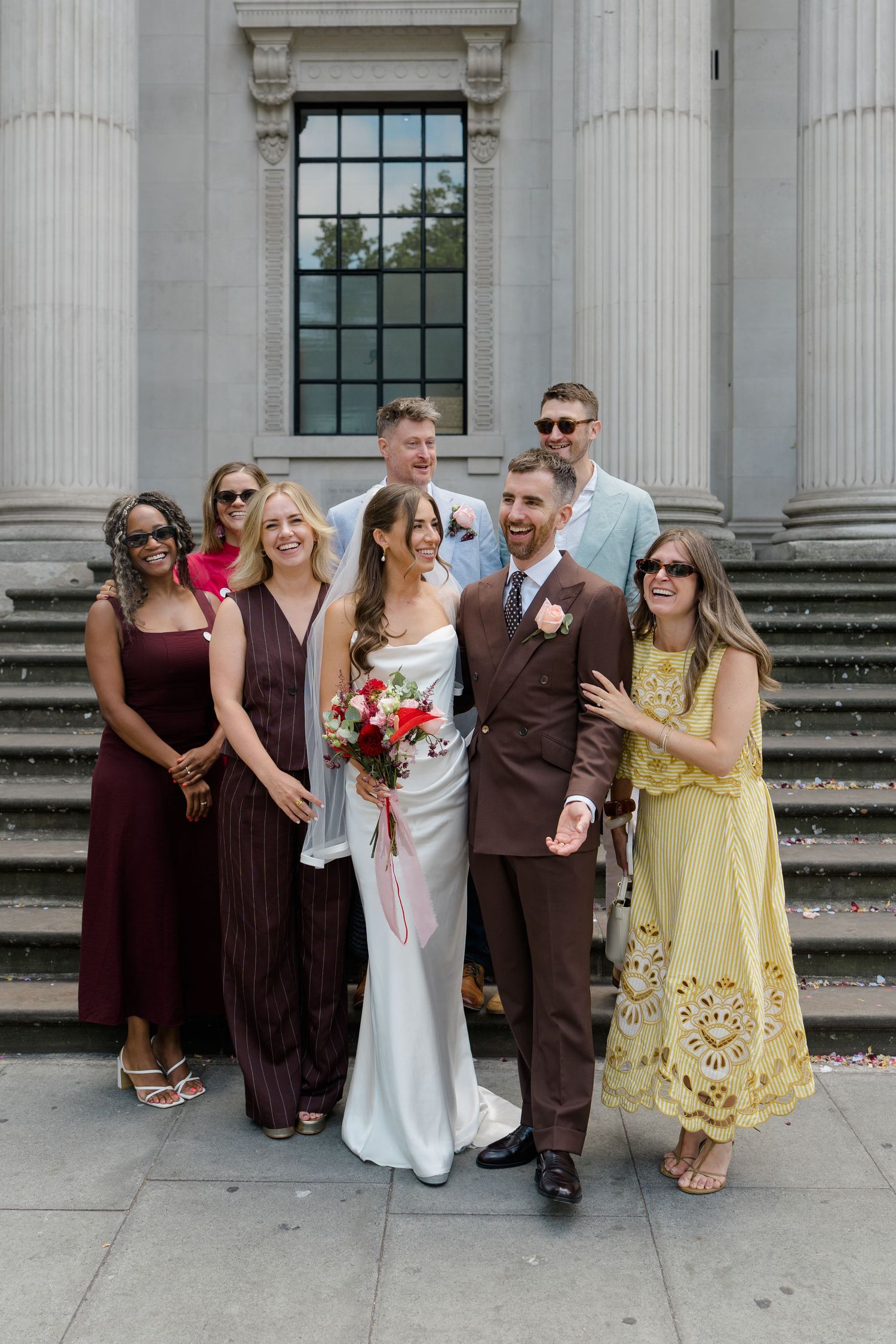 Bride and groom pose with wedding guests on steps of grand stone building on sunny wedding day.