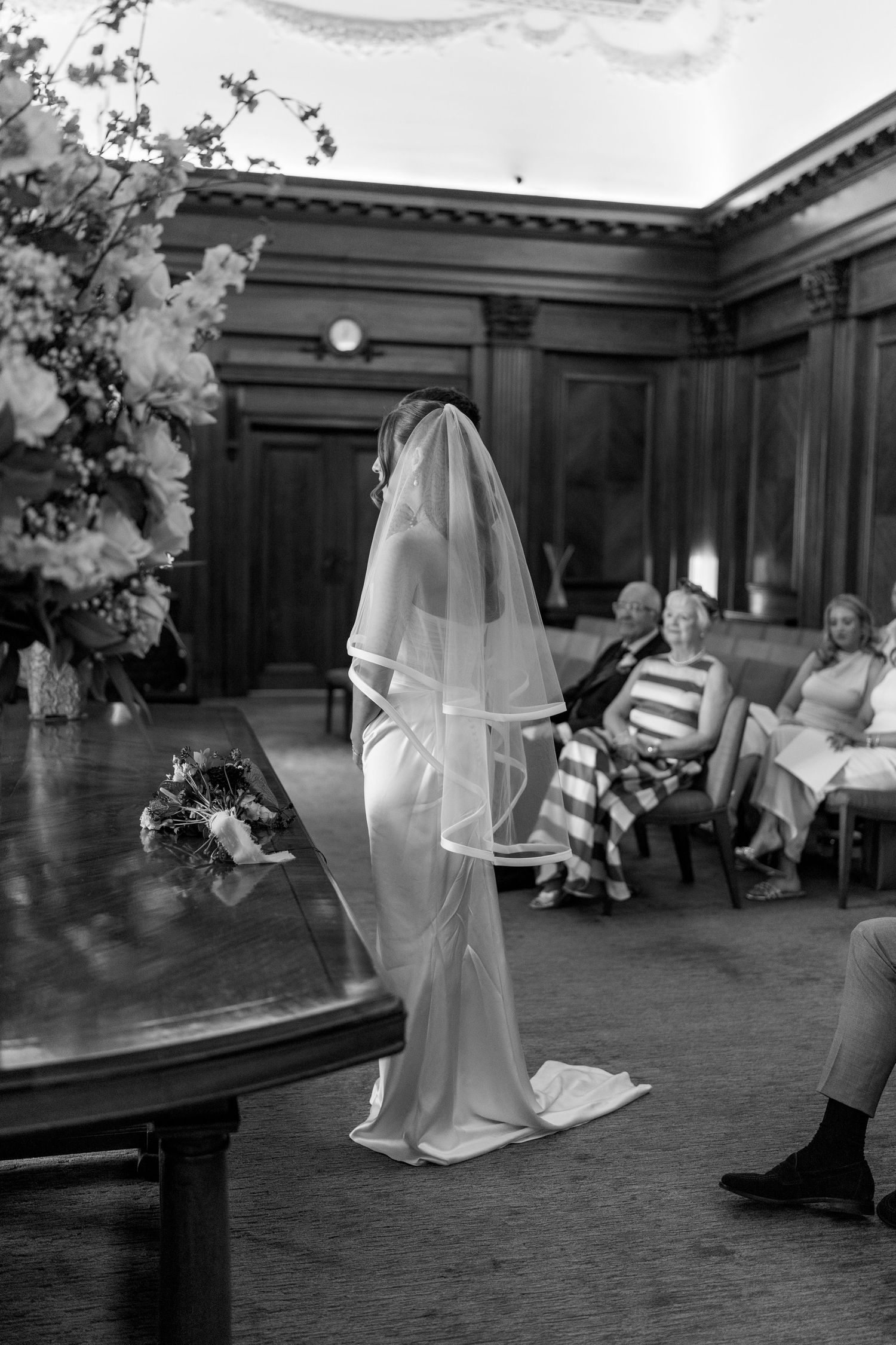 Black and white photo of bride in veil and satin gown standing at ceremony table before seated guests.