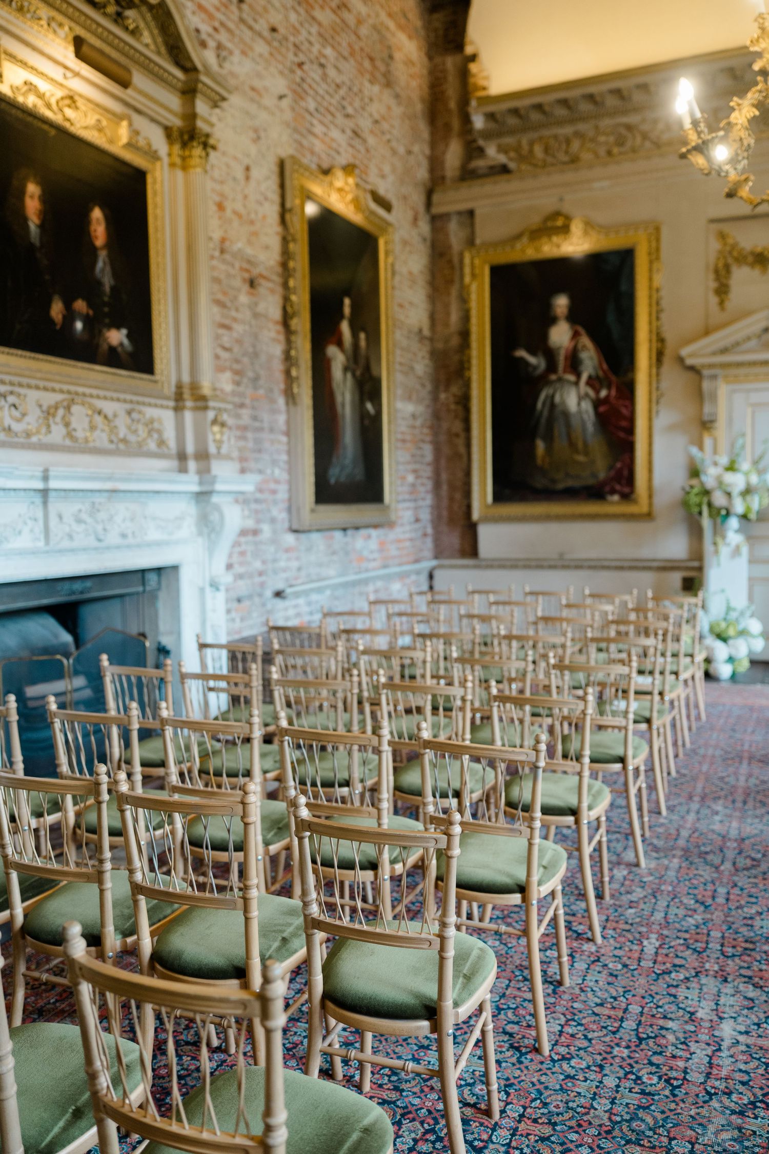 Elegant wedding ceremony setup with gold chiavari chairs, green cushions, ornate portraits, and a marble fireplace in a historic venue.