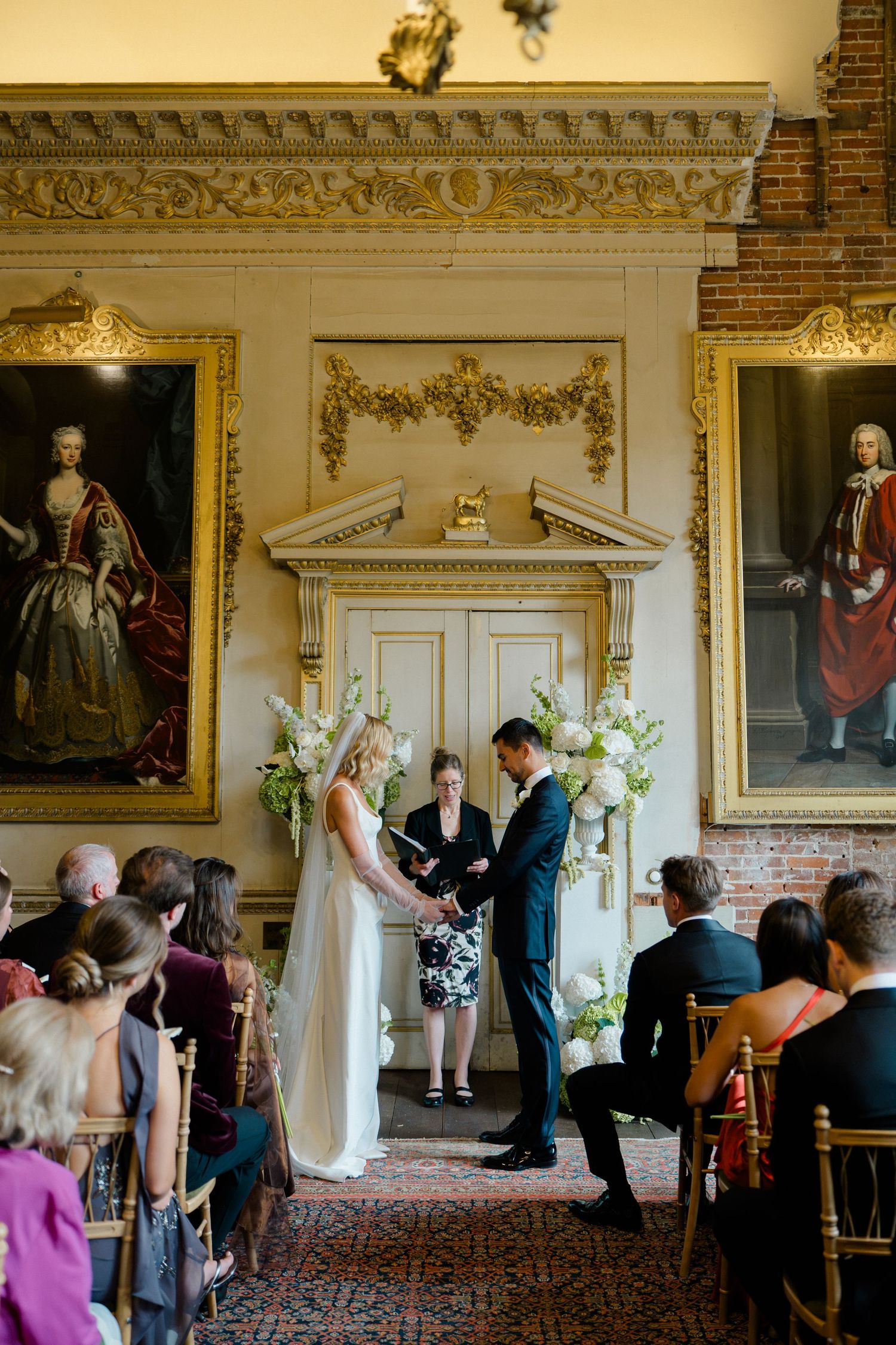 Bride and groom exchange vows in an ornate room with gold-framed portraits and white floral arrangements.