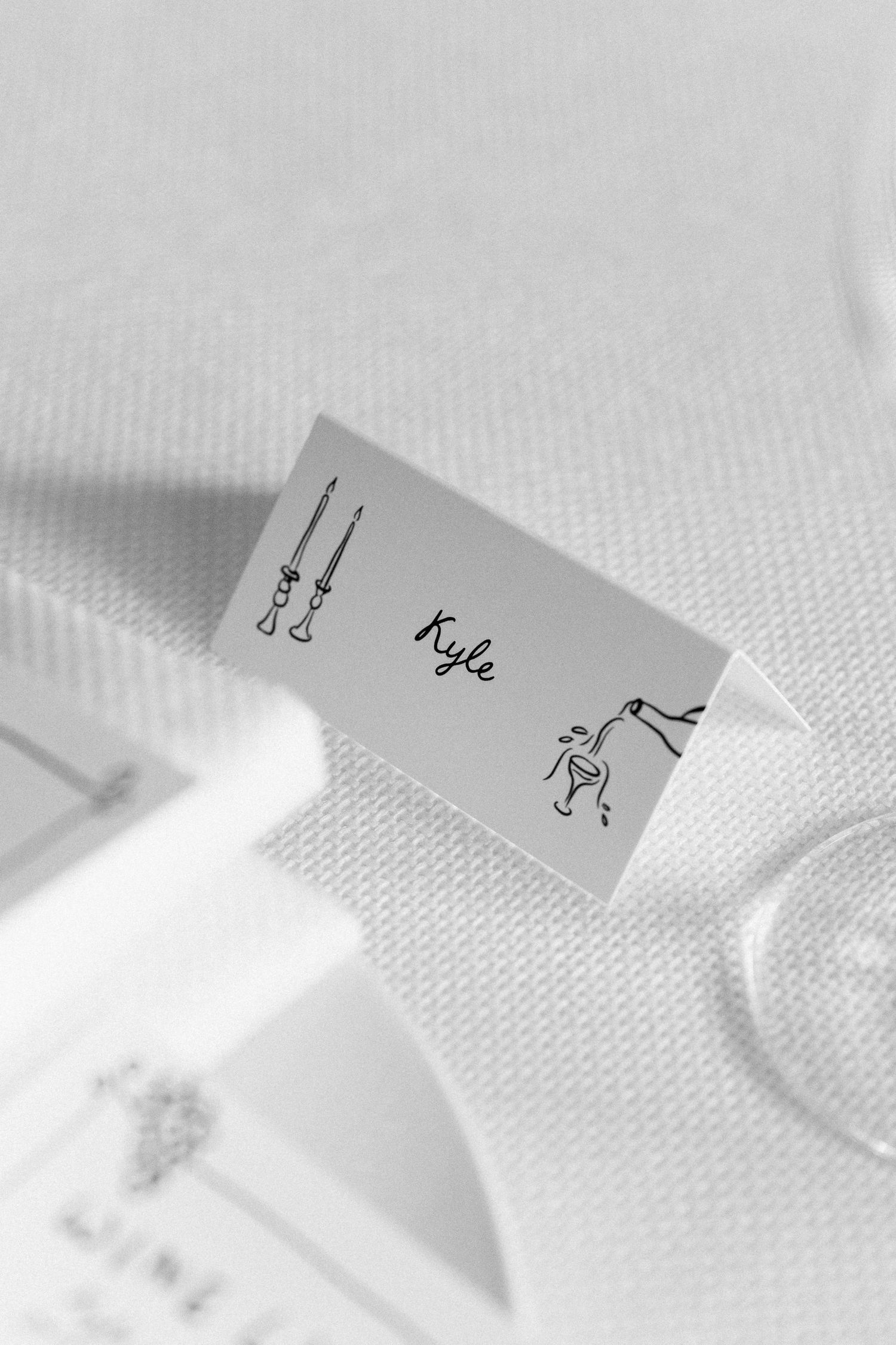 Black and white close-up of a minimalist wedding place card reading Kyle on a white linen table.