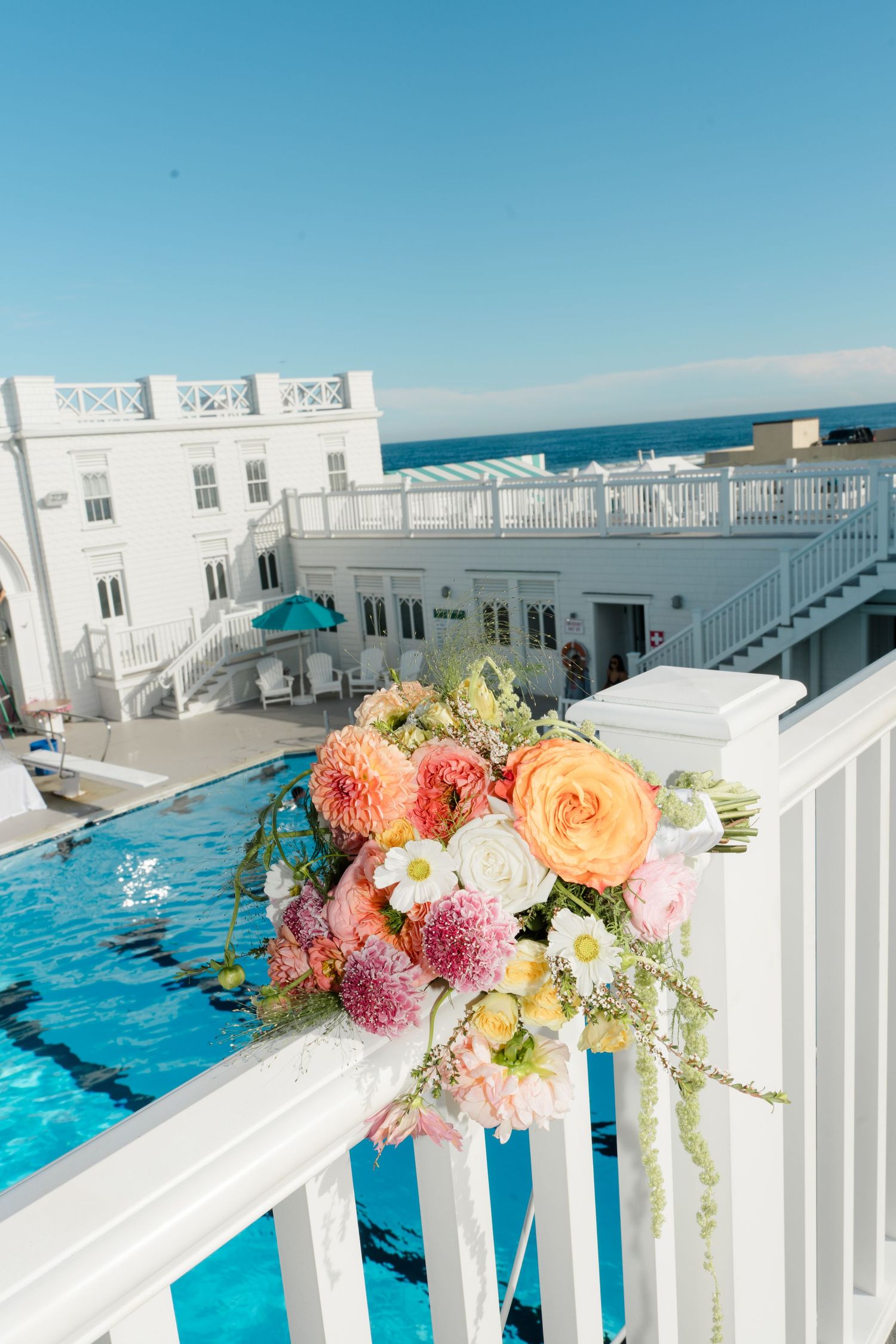 Colorful wedding bouquet with peach roses and pink dahlias draped on white railing overlooking pool and ocean.