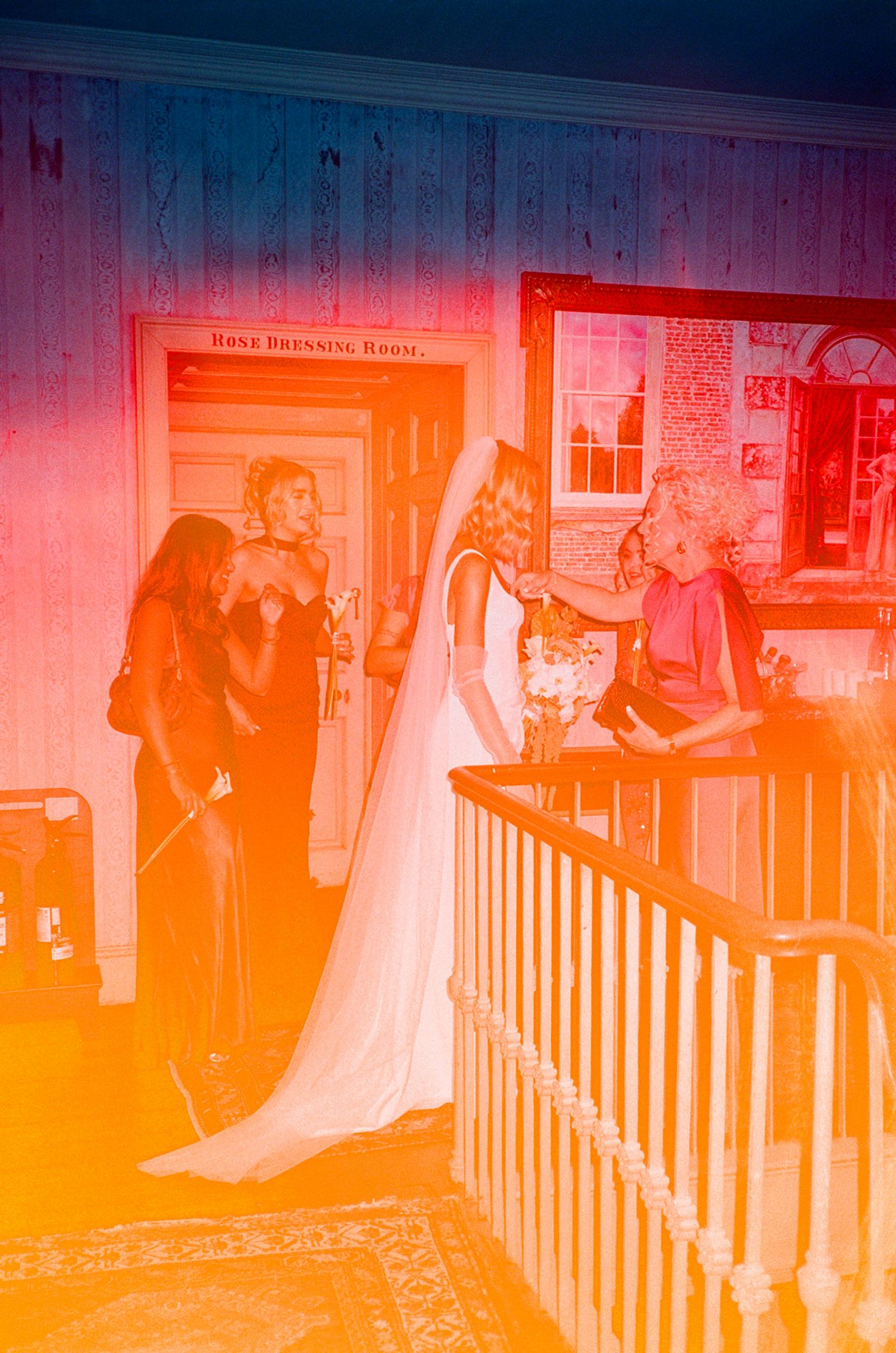 Bride in white gown and veil speaks with guests near a dressing room door in a warmly lit venue.