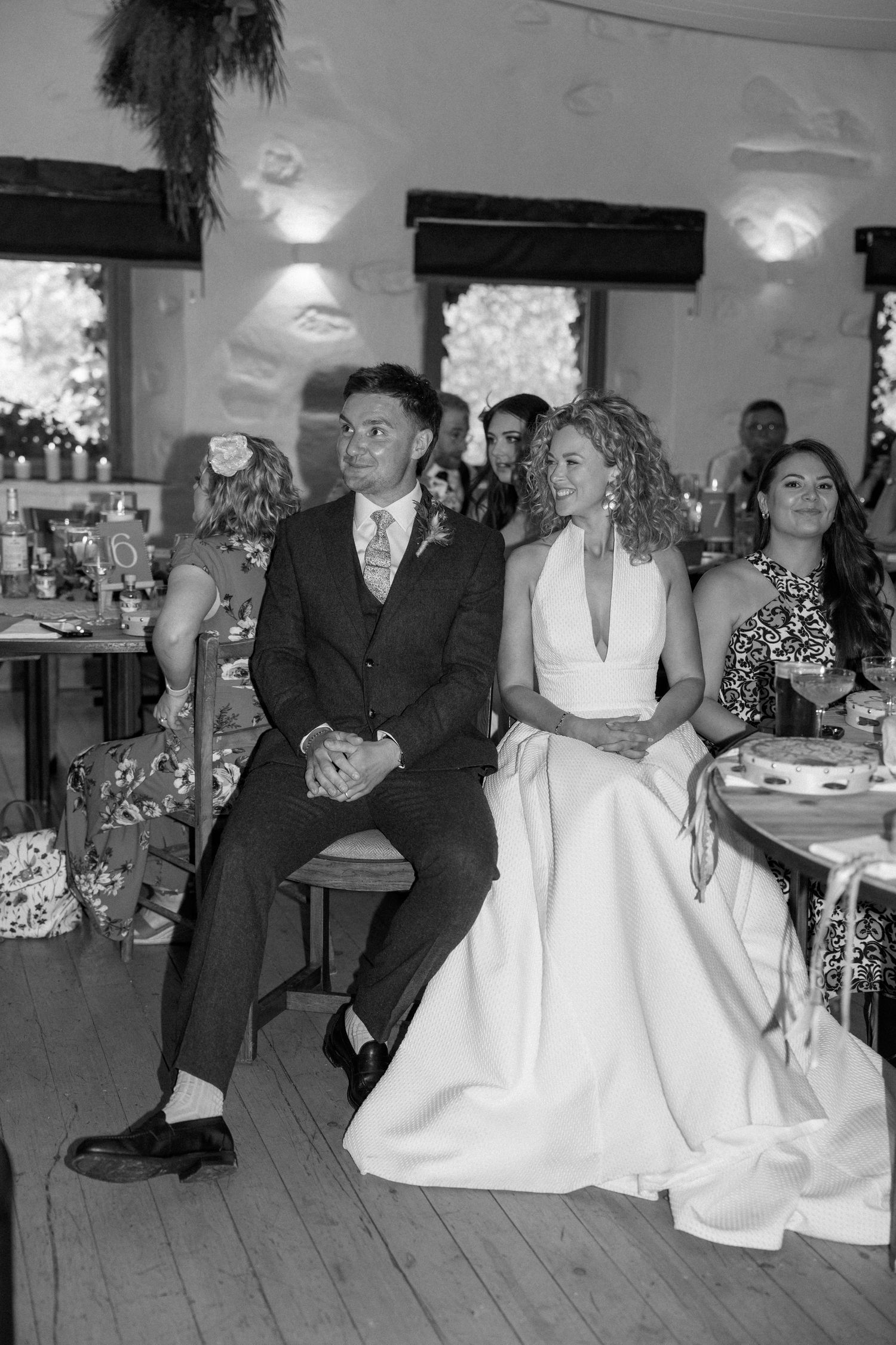 Wedding reception scene with guests seated at tables in rustic venue with white walls.
