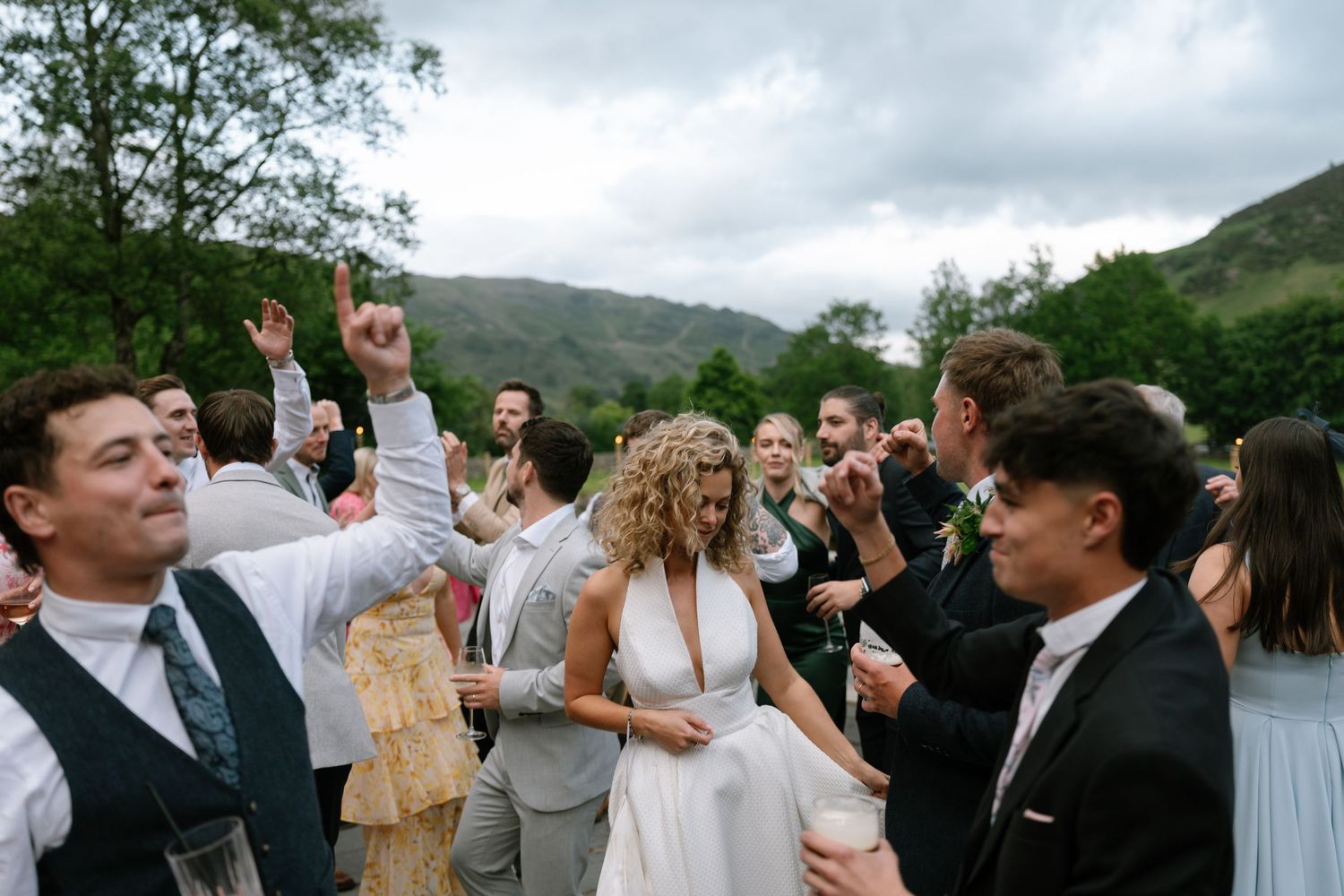 Wedding party celebrates outdoors with mountains and cloudy skies in the background.