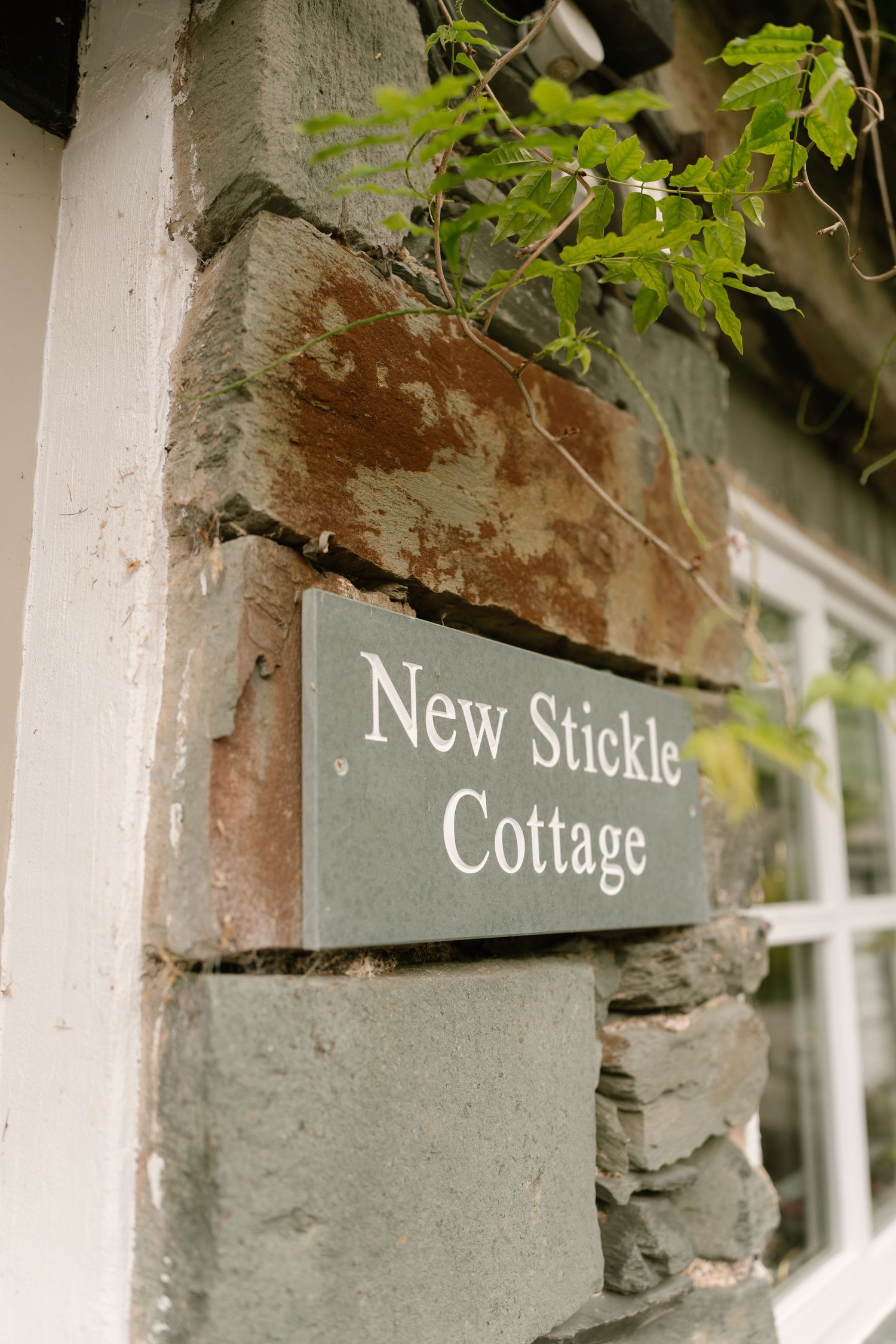 Close-up of a stone cottage sign reading 'New Stickle Cottage' with green foliage above.
