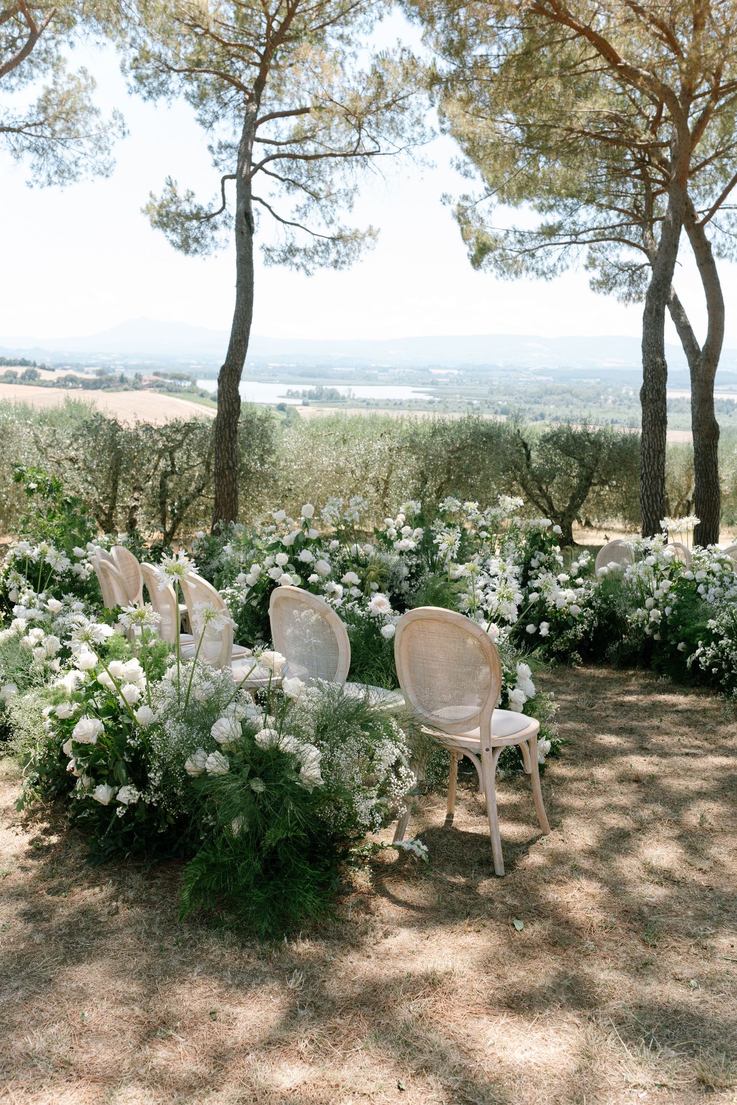 White chairs arranged in rows beneath pine trees with white floral arrangements creating a romantic outdoor ceremony setting.