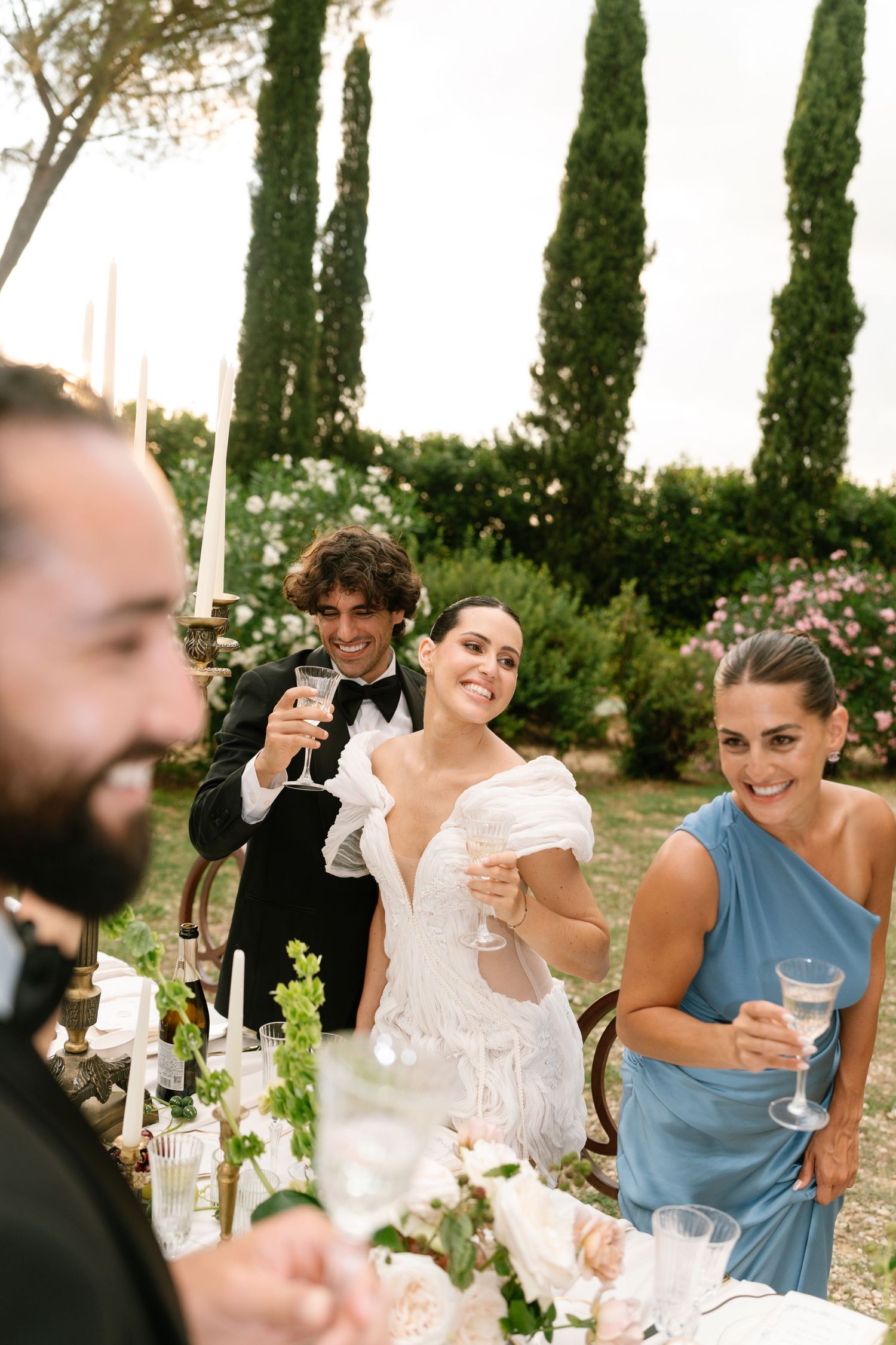 Wedding guests raise glasses in a toast during an outdoor reception among cypress trees.
