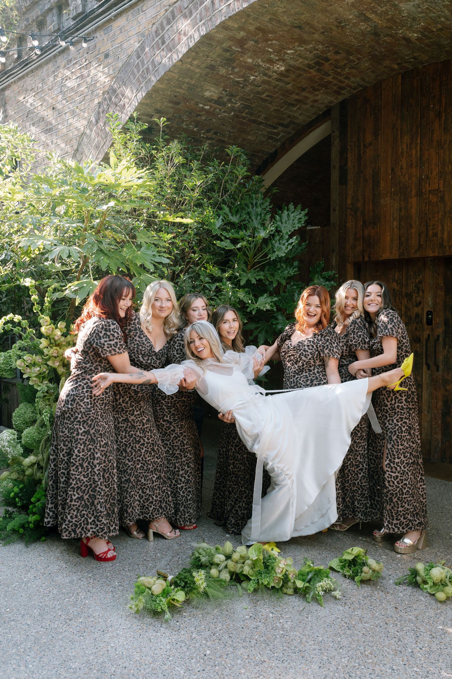 Bridesmaids in matching leopard print dresses pose playfully with the bride in a white gown against a stone wall.
