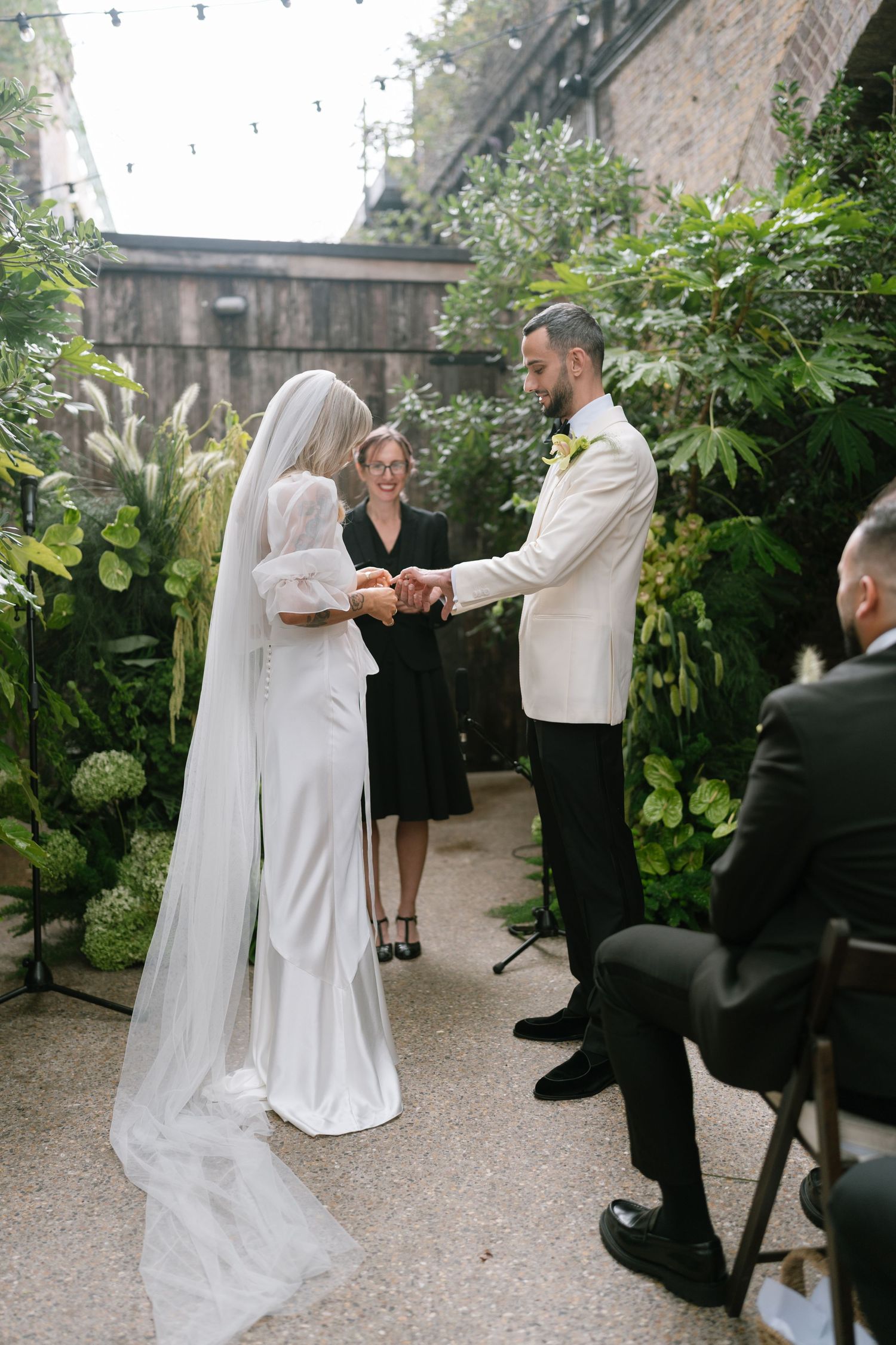 A wedding ceremony unfolds in a lush garden setting with string lights overhead.