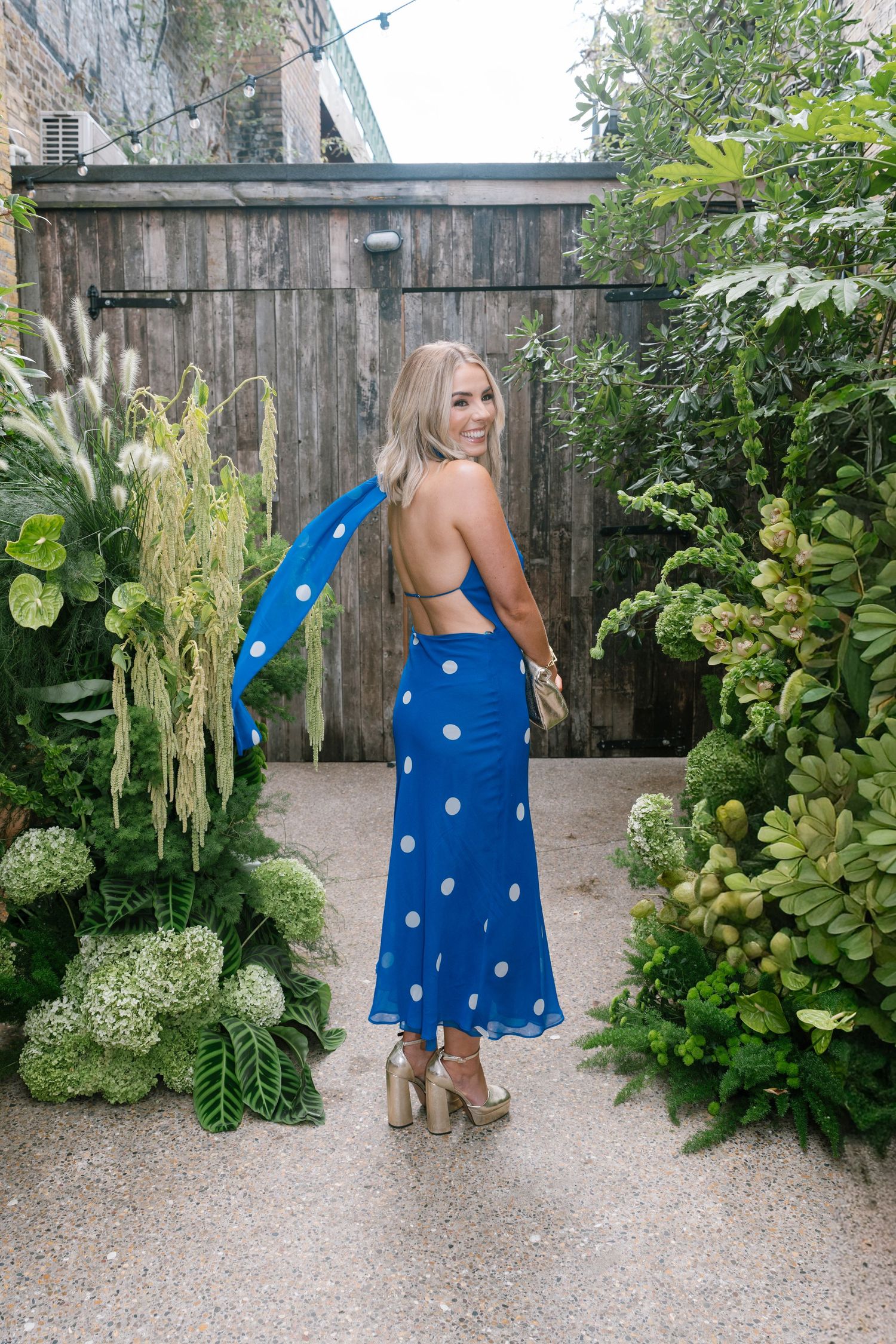 Woman in blue polka dot dress poses in garden setting with lush greenery.