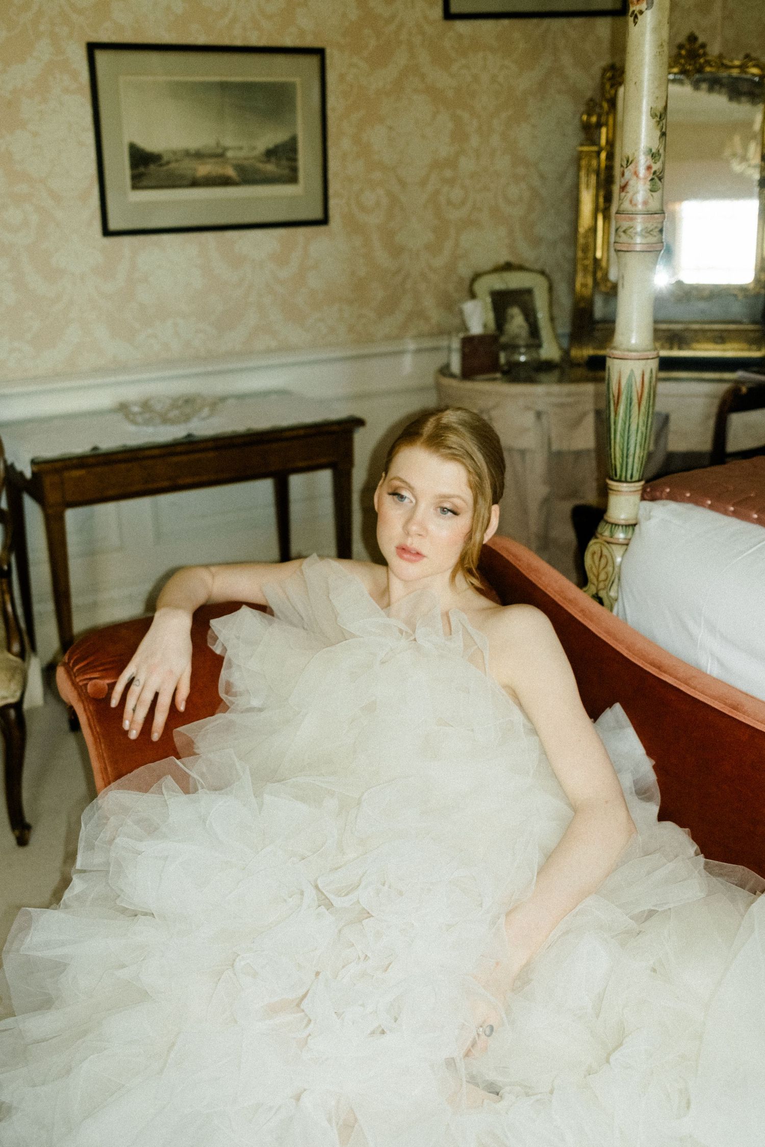 A person in a flowing white tulle gown reclines on an elegant red sofa in a vintage-styled room with floral wallpaper.