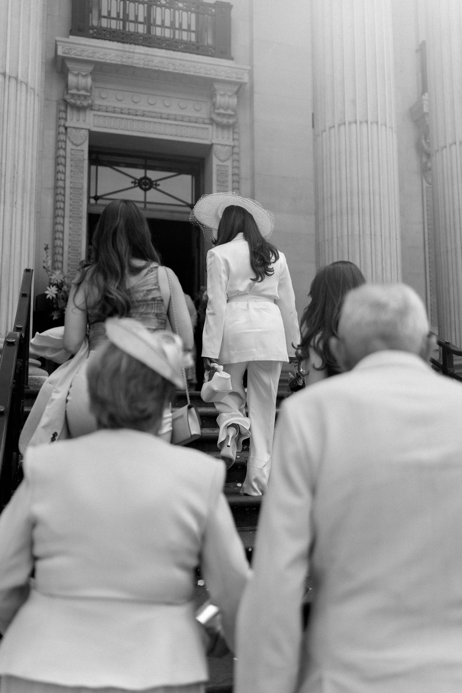 Wedding party ascending grand columned steps, bride in white pantsuit and hat leading the group.