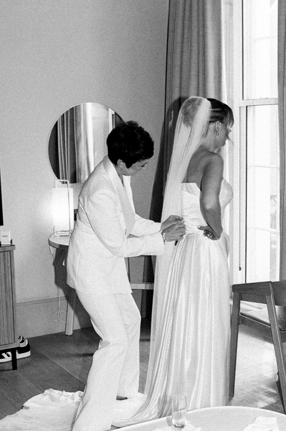 Black and white photo of a person helping a bride in a white gown and veil prepare near a window.