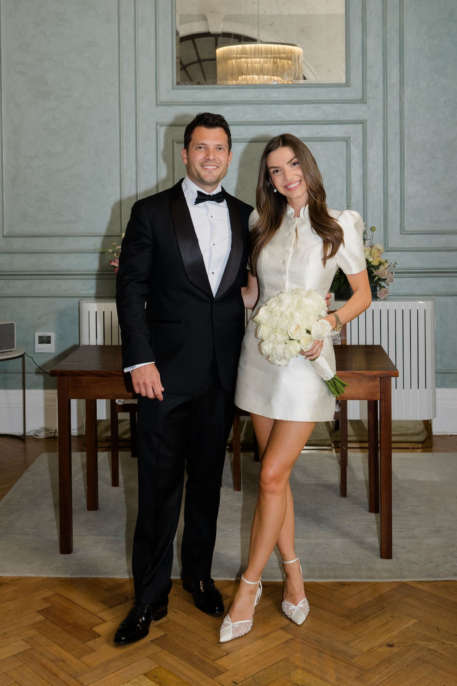 Newlyweds pose together in elegant room, groom in tuxedo and bride in white mini dress with bouquet.