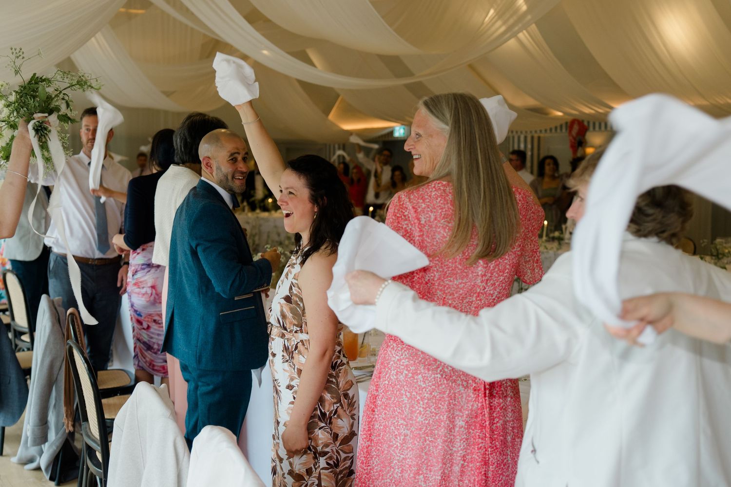 Wedding guests wave white napkins in celebration inside an elegant marquee reception venue.
