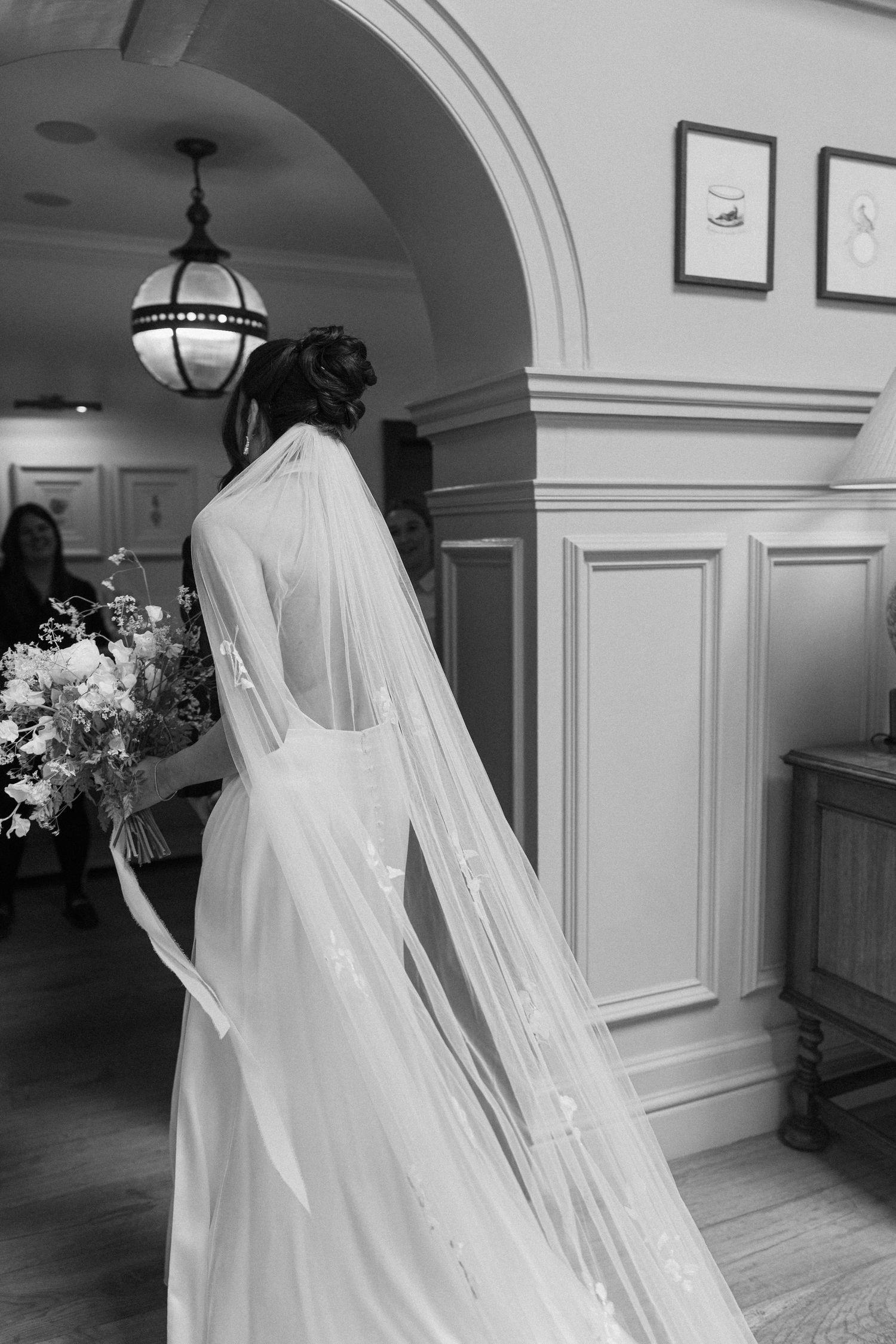Black and white photo of bride in elegant gown with long lace veil holding floral bouquet indoors.
