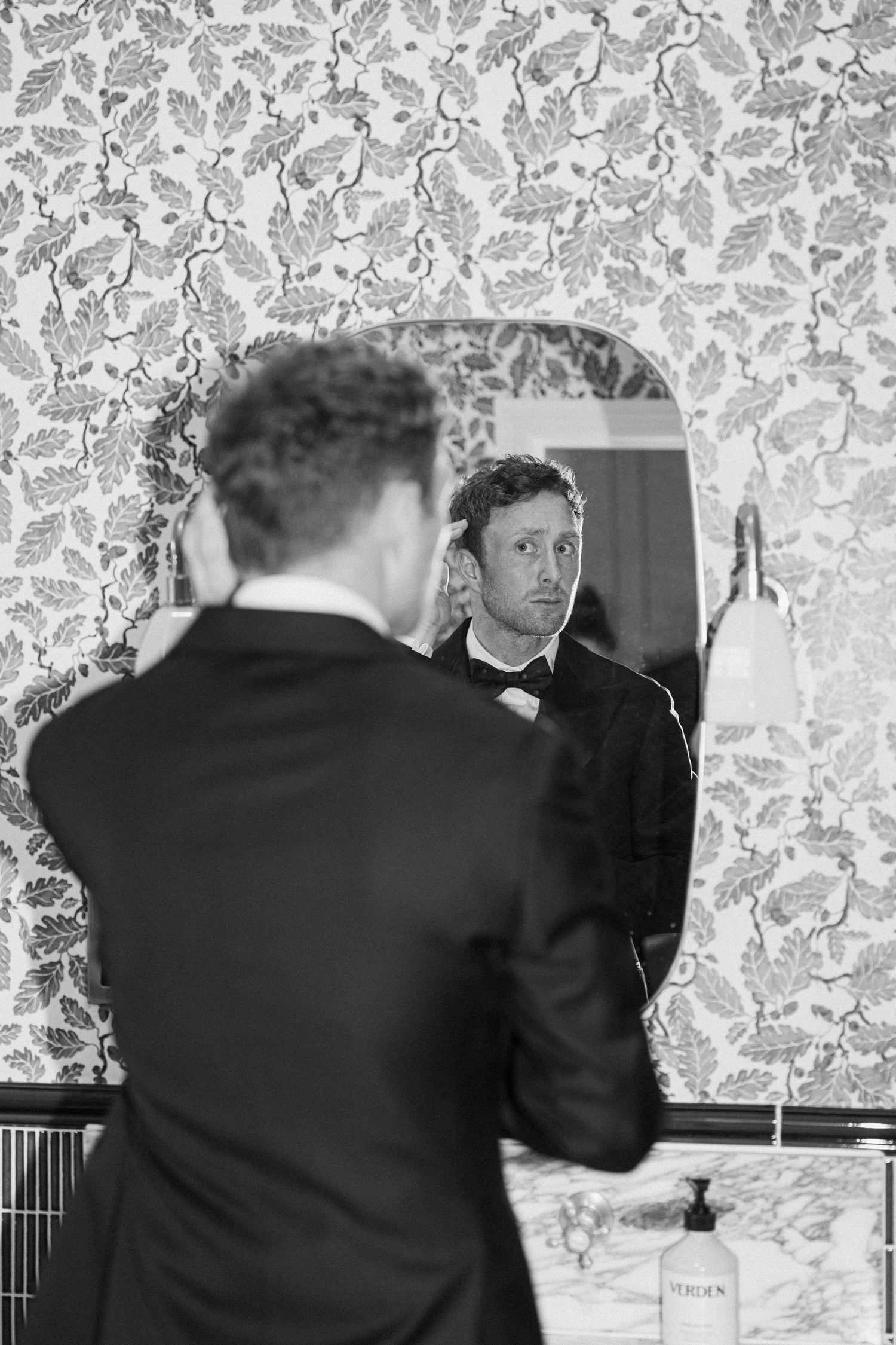 Groom in black tuxedo examining his reflection in an ornate bathroom mirror with floral wallpaper.