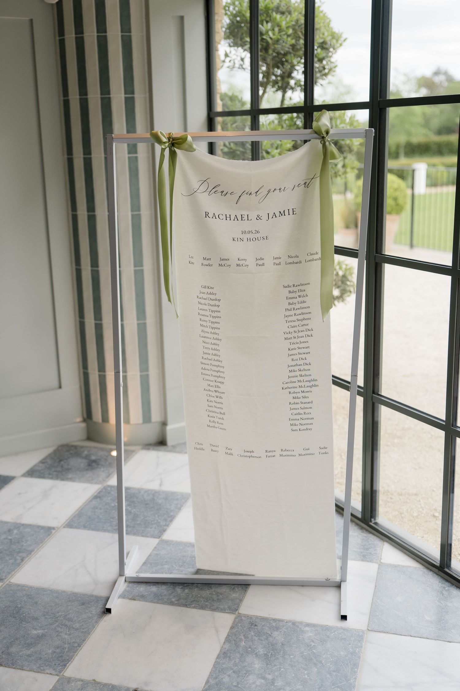 Elegant linen seating chart for Rachael & Jamie displayed on metal stand by large window.