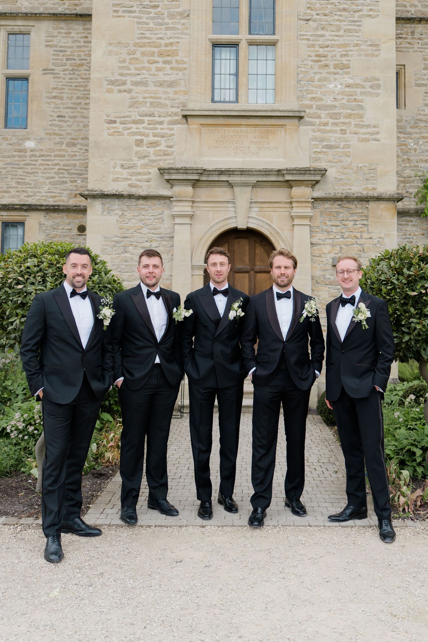 Five groomsmen in matching black tuxedos with boutonnieres pose before stone manor entrance.