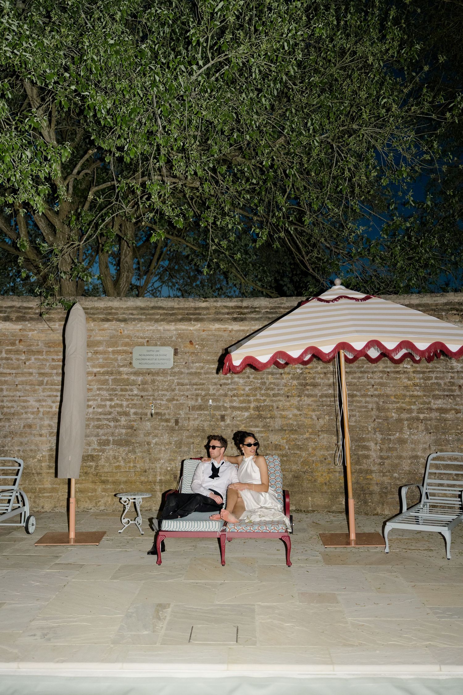 Couple in formal attire sitting together under a striped umbrella by a pool at night.