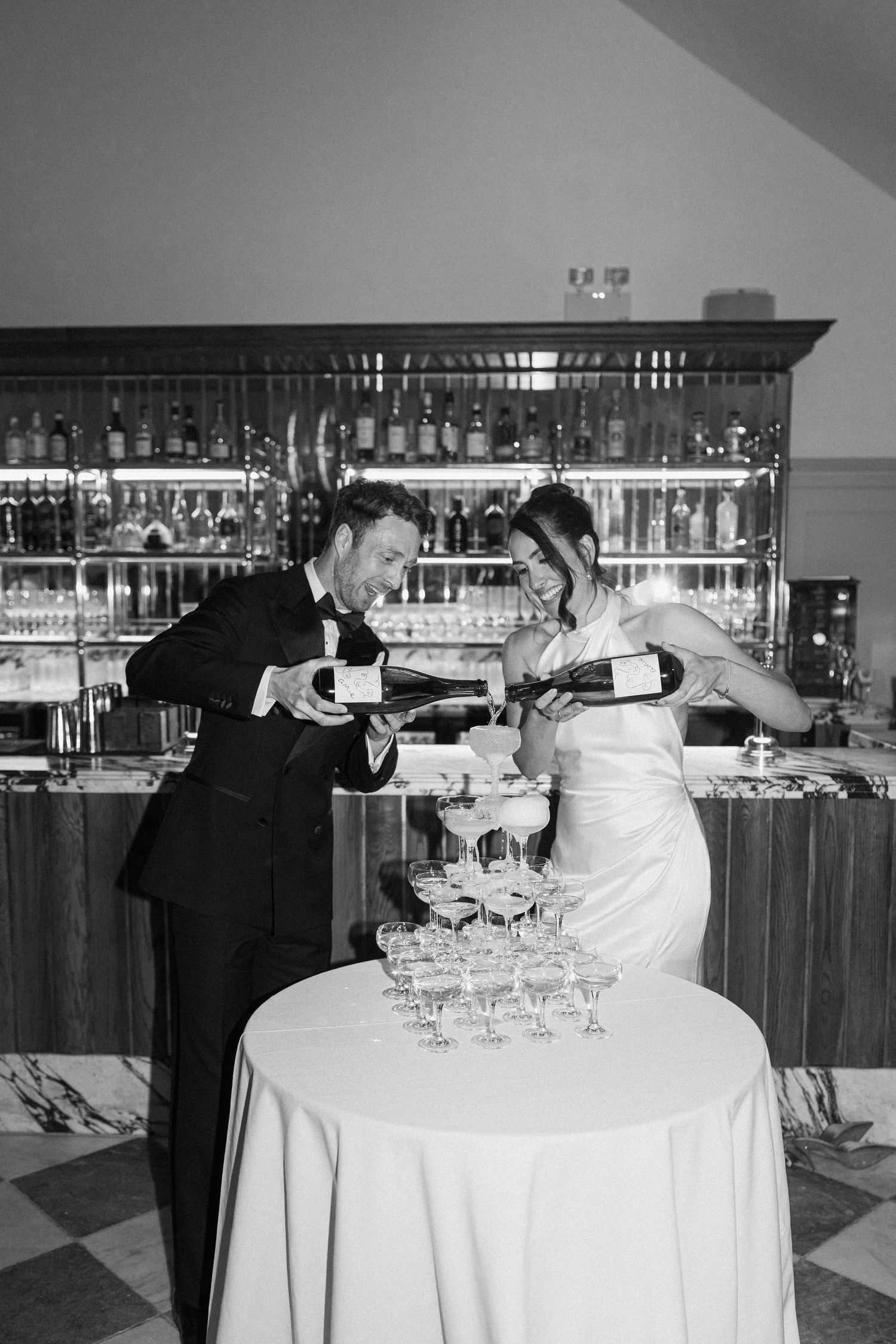 Newlyweds laughing while pouring champagne tower at elegant wedding reception.